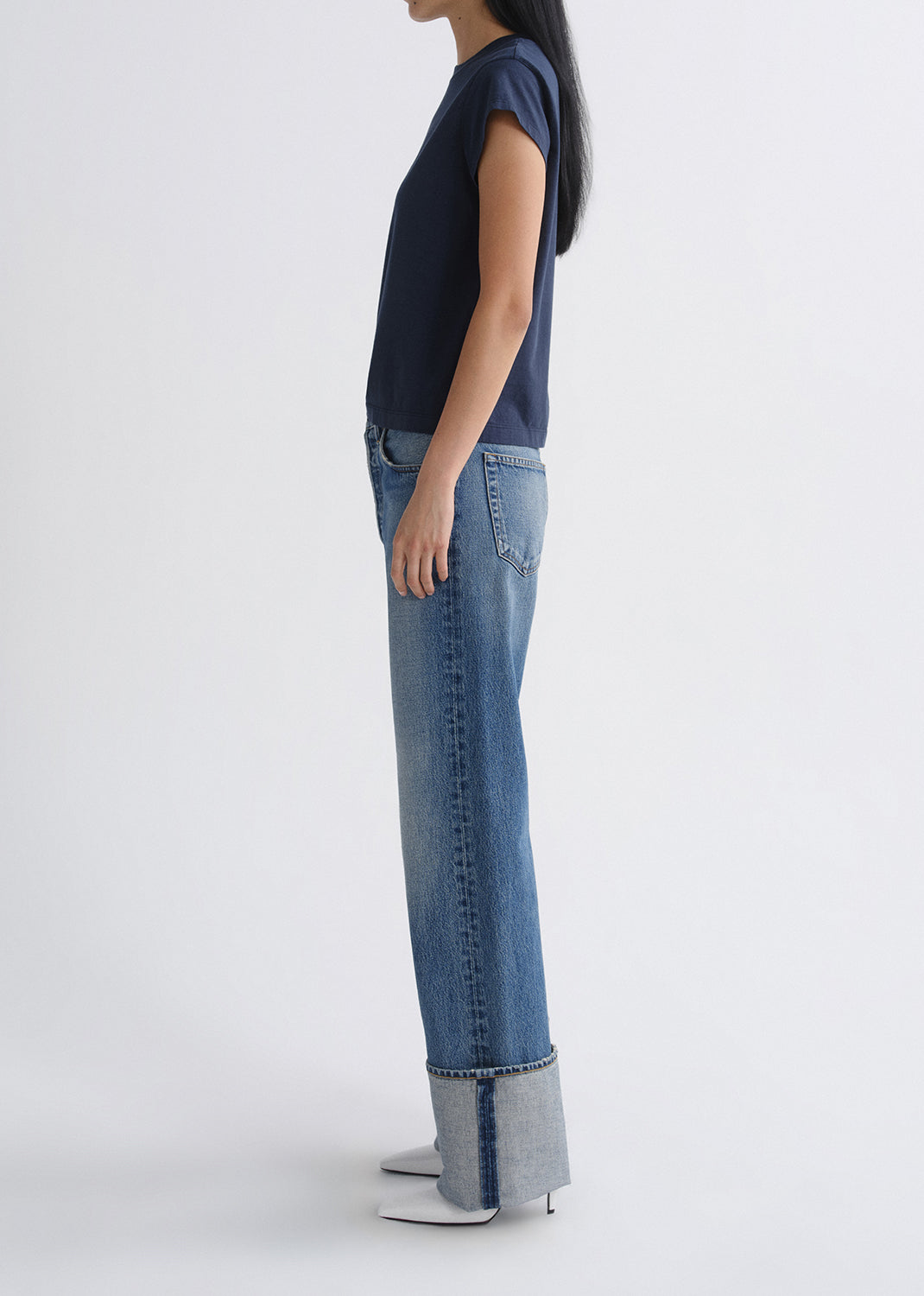 A model shown from the side profile wearing a navy blue short-sleeve top, light-wash blue jeans with a rolled hem, and white pointed-toe heels against a grey background.