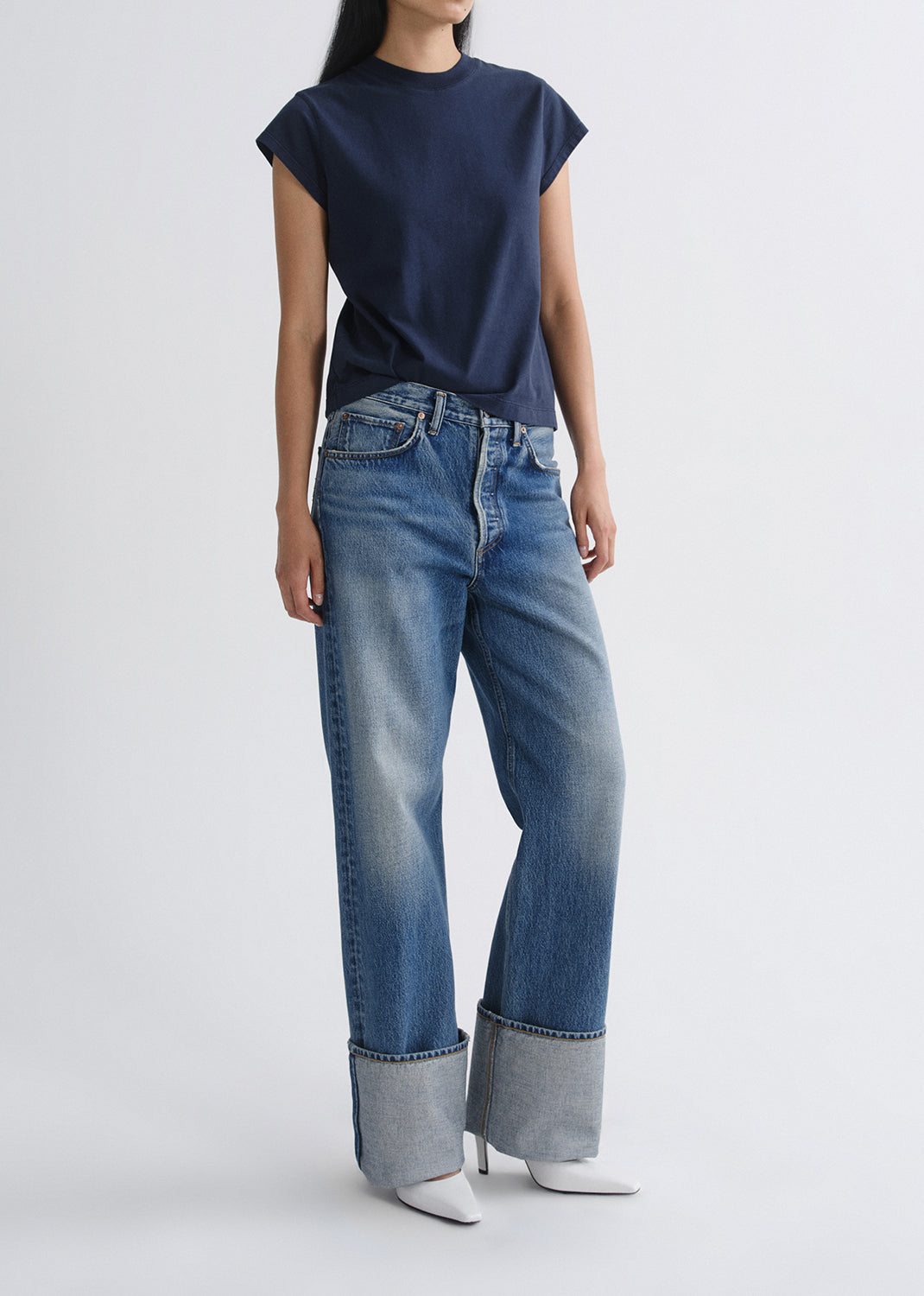 A model wearing a navy blue short-sleeve t-shirt and wide-leg blue jeans with rolled cuffs, paired with white pointed-toe heels, against a grey background.