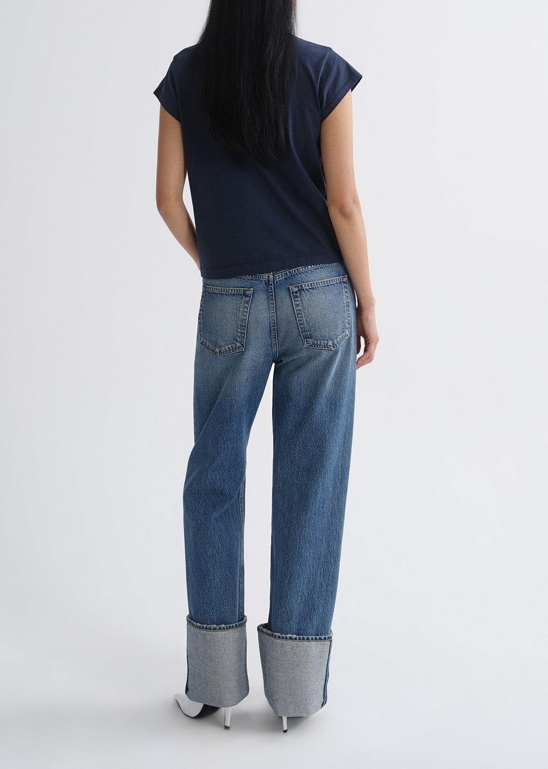 A model shown from the back wearing a navy blue t-shirt and wide-leg blue jeans with rolled cuffs, set against a grey background.