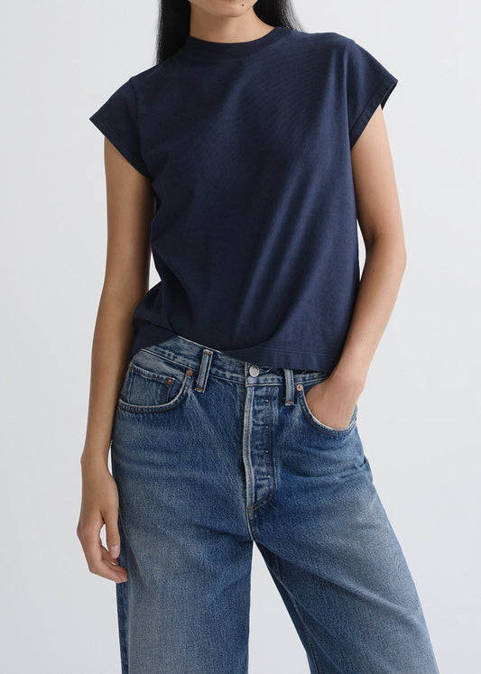 A person wearing a dark navy blue short-sleeve crewneck t-shirt tucked into blue jeans, shown from the neck down against a grey background.