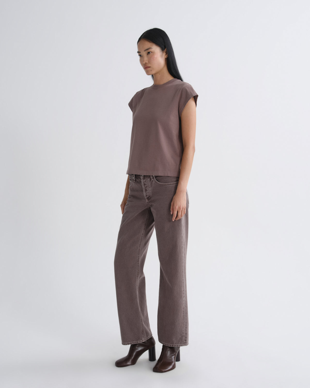 A woman modeling a muted mauve-brown short-sleeve top and matching wide-leg pants with dark brown heeled boots, standing against a plain light gray studio background.