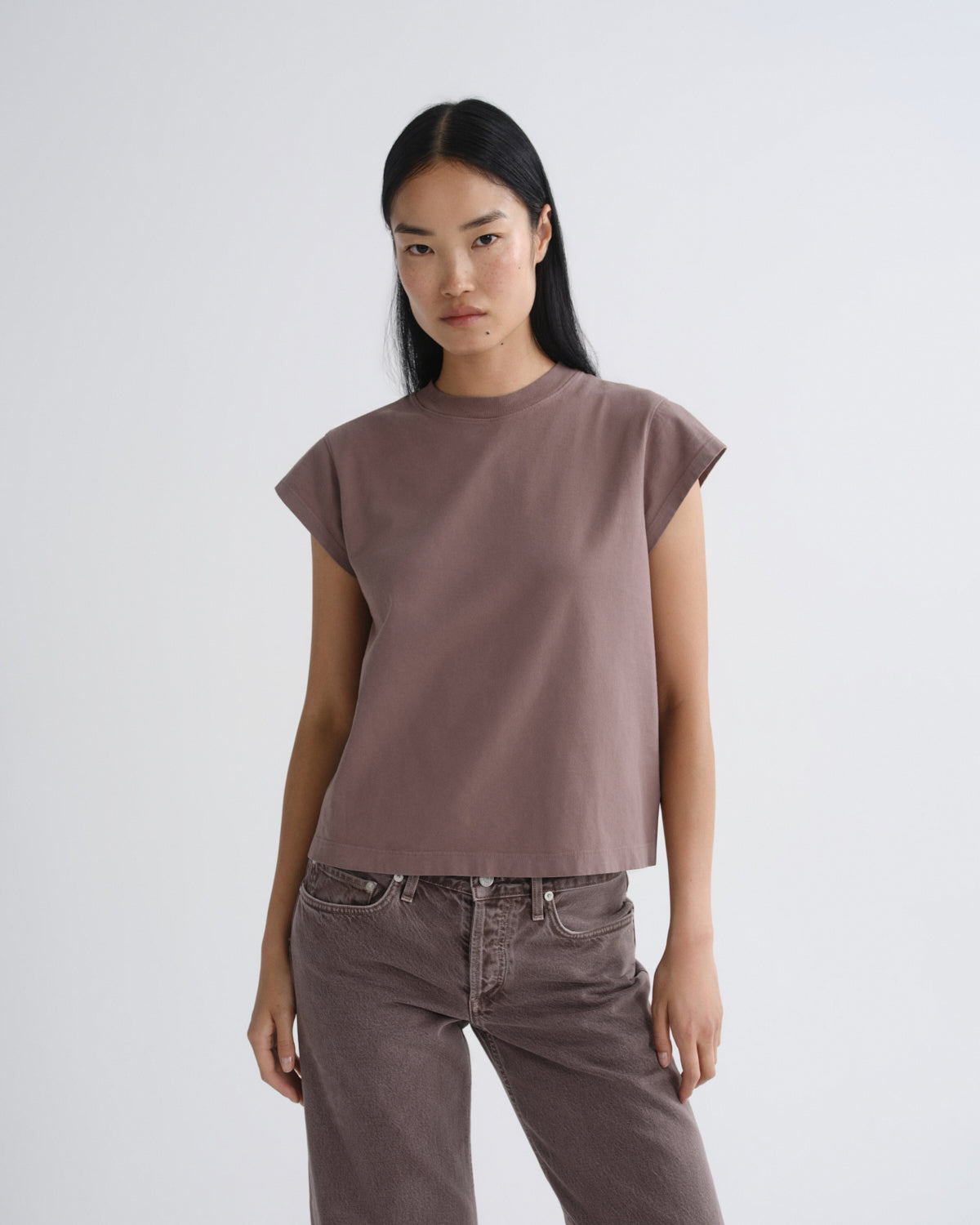 A woman modeling a muted mauve-brown short-sleeve crop top with a relaxed fit, paired with dark brown pants, against a plain light gray studio background.