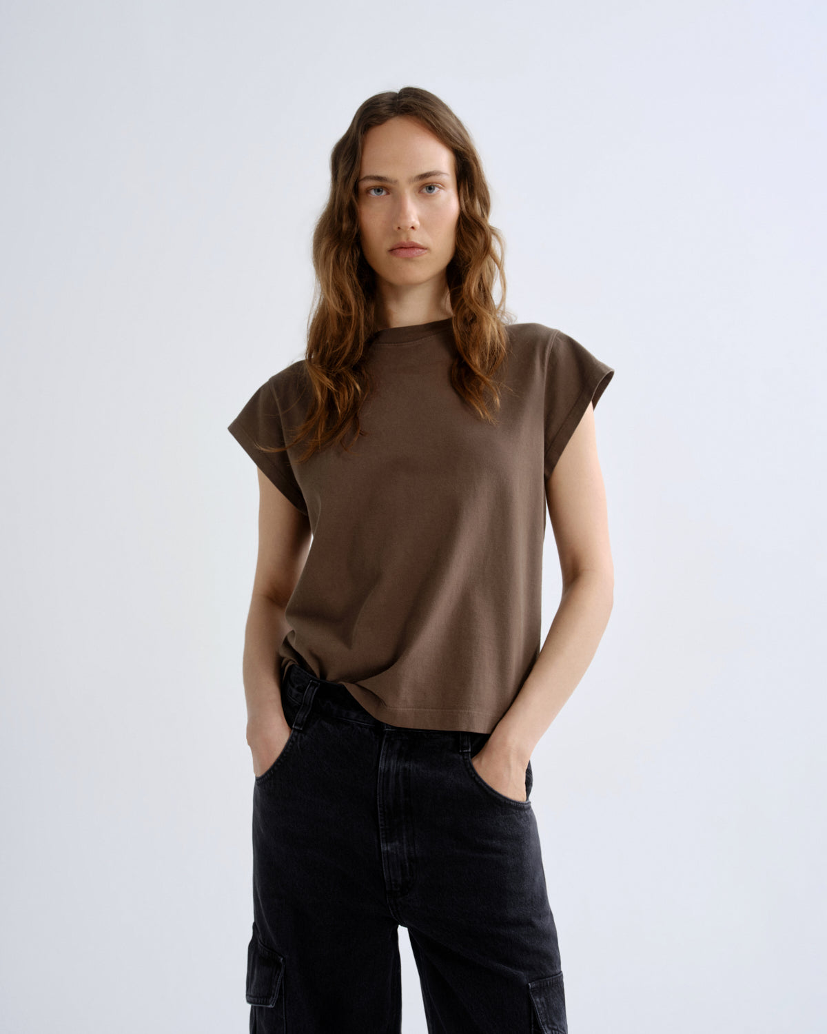 A woman modeling a brown short-sleeve crewneck t-shirt with black cargo pants, standing against a light gray studio background.