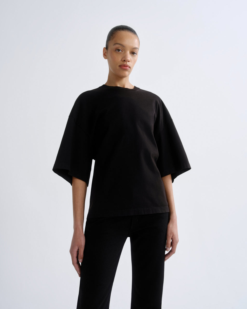 A model wearing a loose-fitting black short-sleeve t-shirt and black pants, standing against a light gray studio background.
