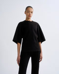 A model wearing a loose-fitting black short-sleeve t-shirt and black pants, standing against a light gray studio background.