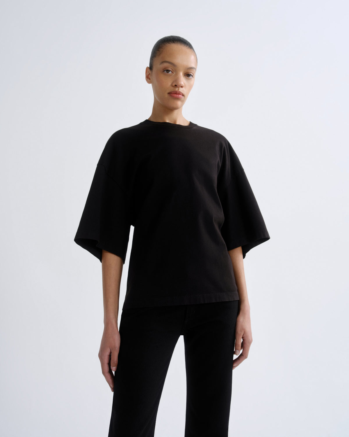 A model wearing a loose-fitting black short-sleeve t-shirt and black pants, standing against a light gray studio background.