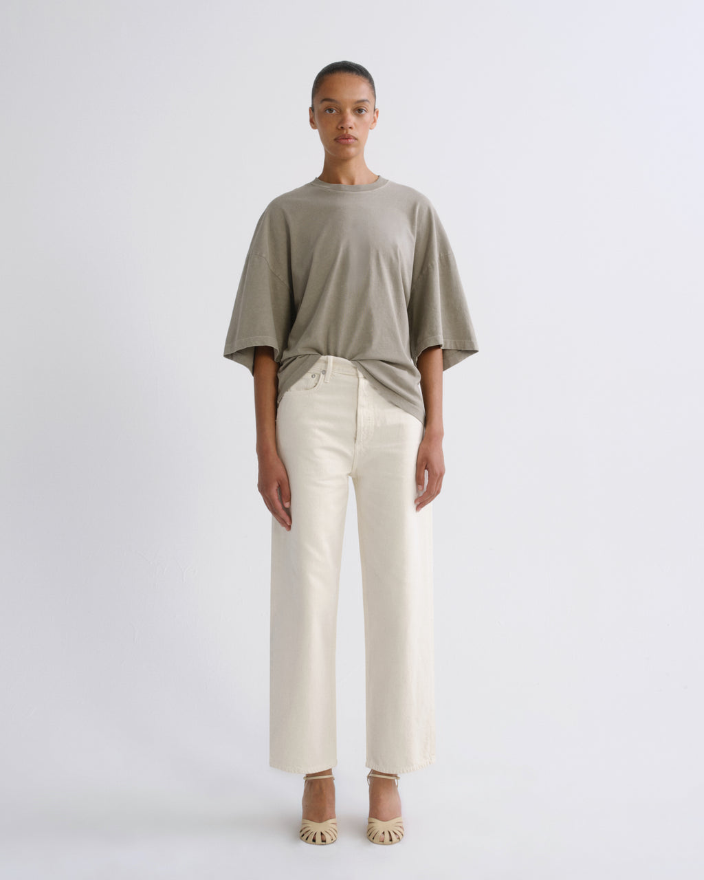 A model stands against a light gray studio background wearing an oversized taupe short-sleeve t-shirt tucked into off-white wide-leg cropped trousers, paired with beige pointed-toe heels.