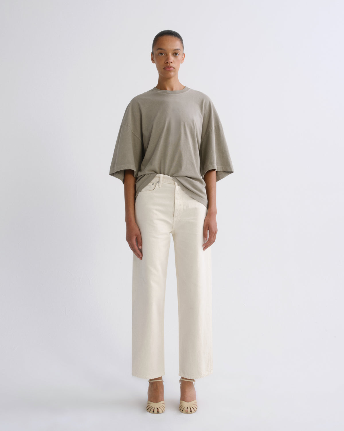 A model stands against a light gray studio background wearing an oversized taupe short-sleeve t-shirt tucked into off-white wide-leg cropped trousers, paired with beige pointed-toe heels.