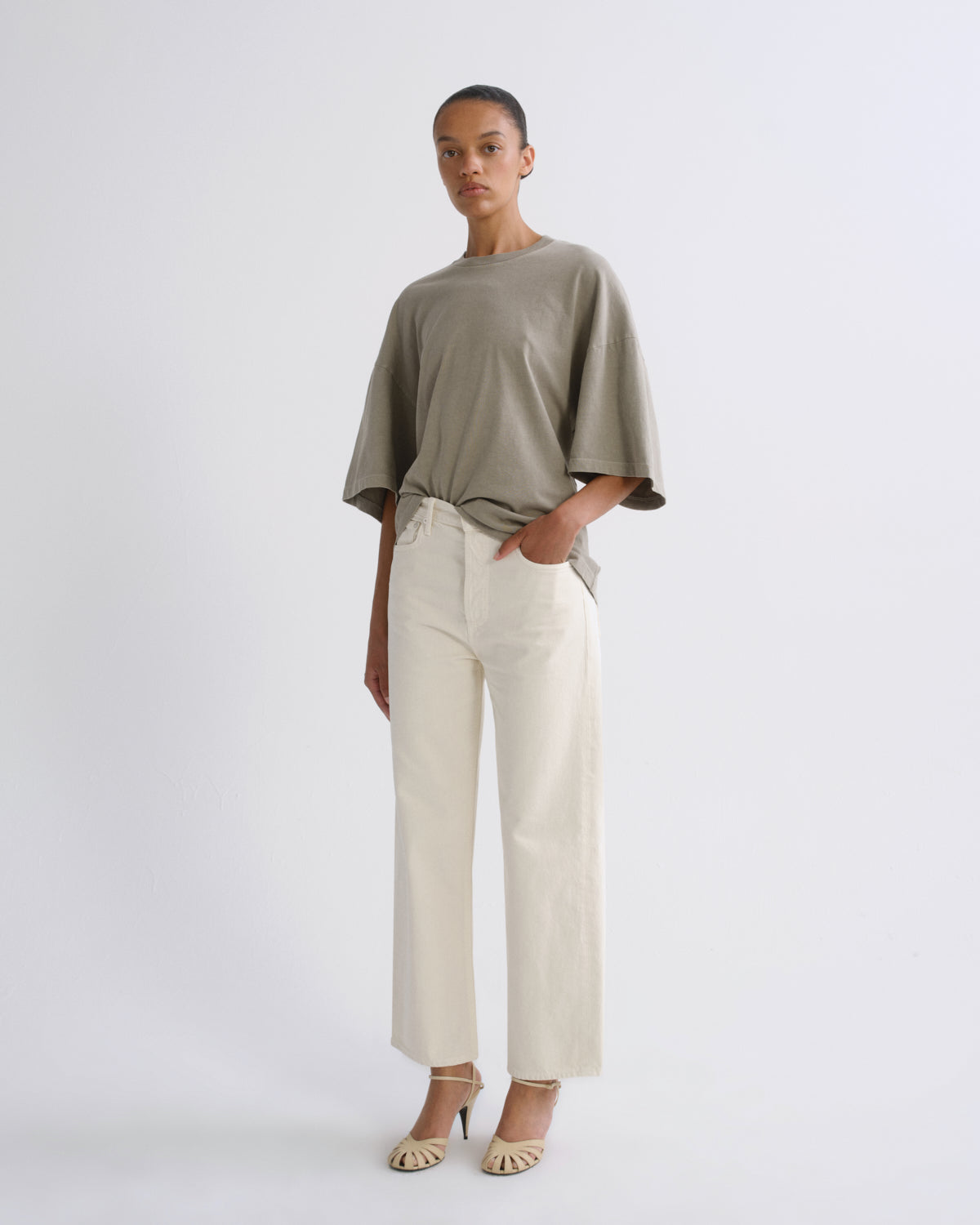 A model stands against a light gray studio background wearing an oversized taupe-green short-sleeve top, off-white wide-leg cropped trousers, and beige heeled sandals.