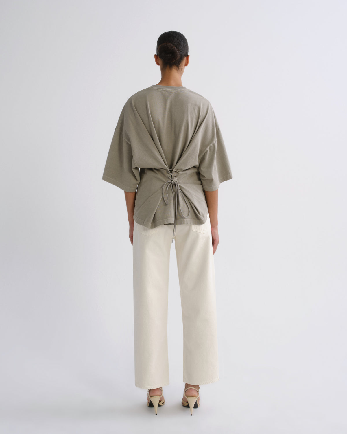 A model viewed from the back wearing a light olive green oversized short-sleeve top with a knotted waist detail, paired with off-white wide-leg trousers and beige heels, against a plain light gray studio background.