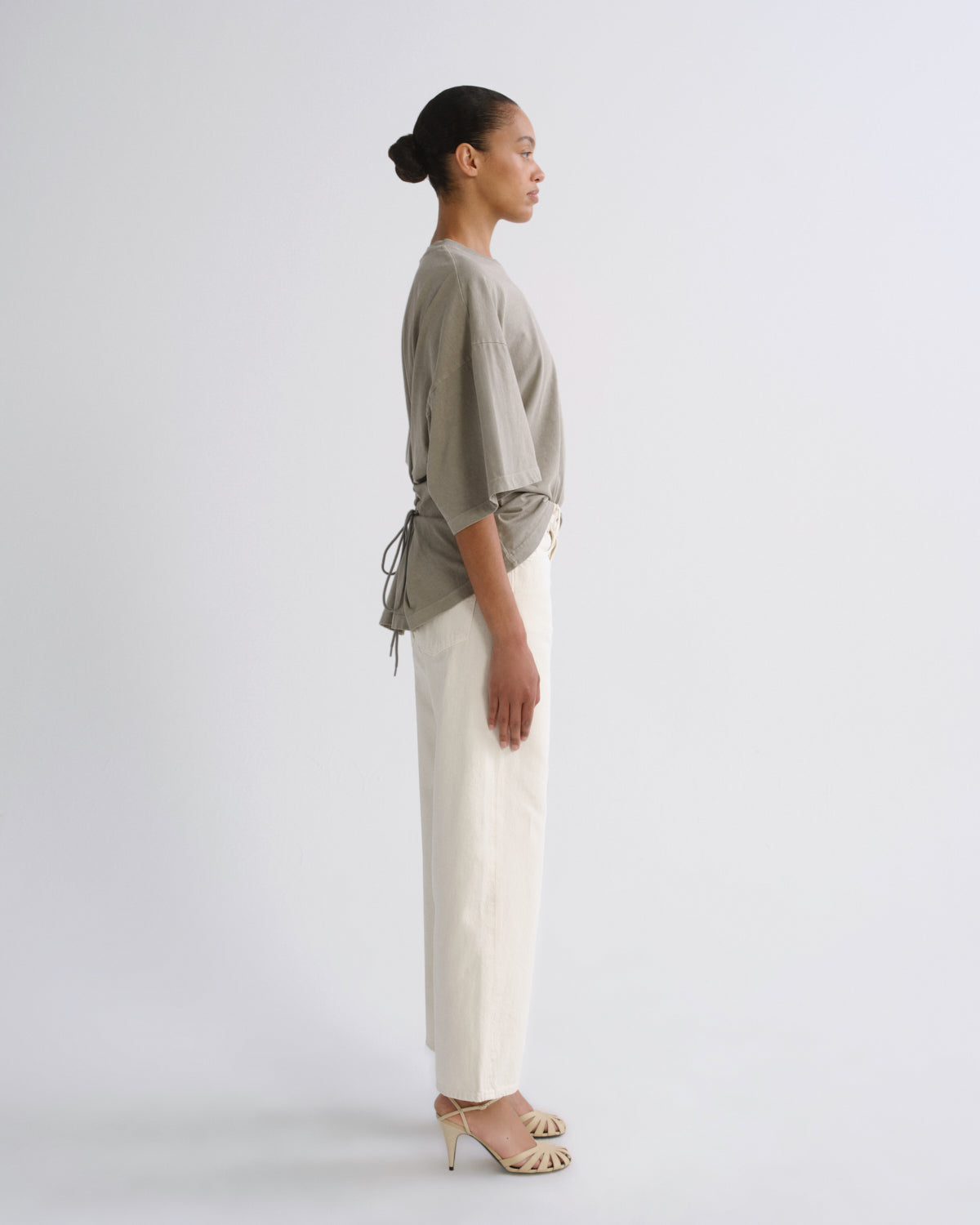 A model is shown in profile wearing a light olive green short-sleeve top with side drawstrings and cream wide-leg trousers, paired with beige high-heeled sandals, against a plain light gray studio background.