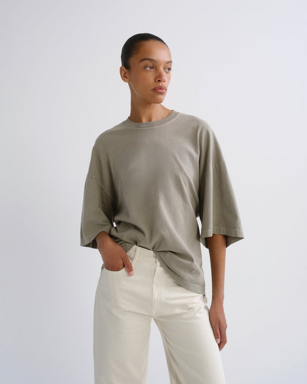 A model wearing an oversized, short-sleeved crewneck t-shirt in a muted sage green, paired with off-white trousers, standing against a plain light gray studio background.