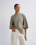 A model wearing an oversized, short-sleeved crewneck t-shirt in a muted sage green, paired with off-white trousers, standing against a plain light gray studio background.