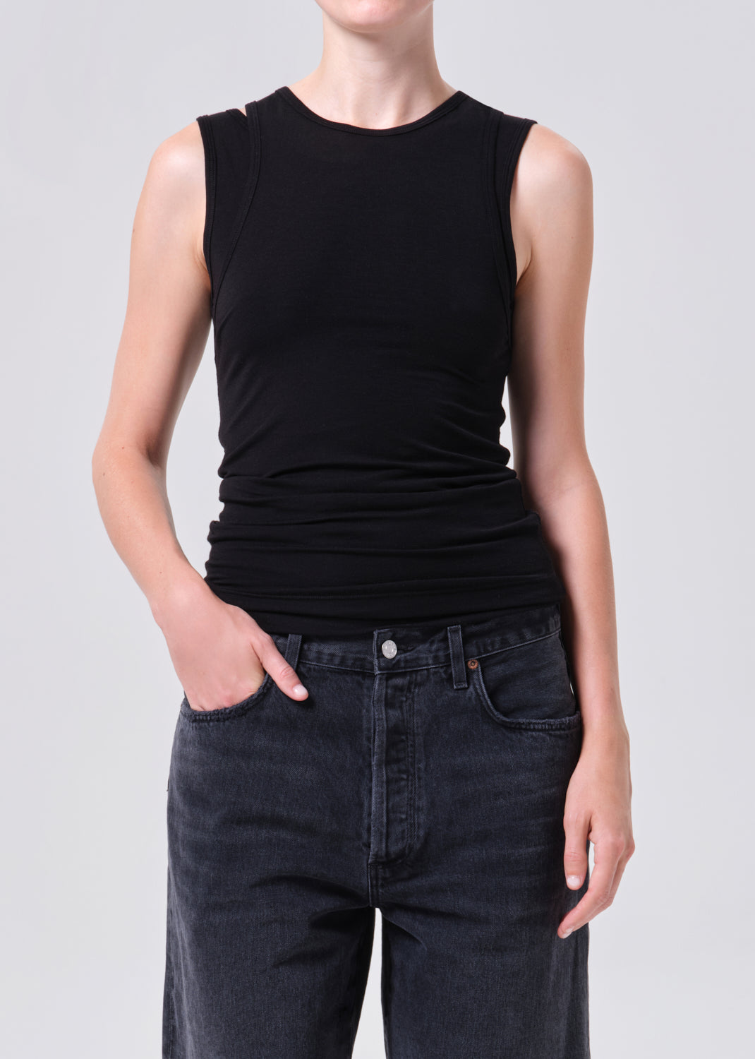 Person wearing a black sleeveless top and dark jeans on a light gray background