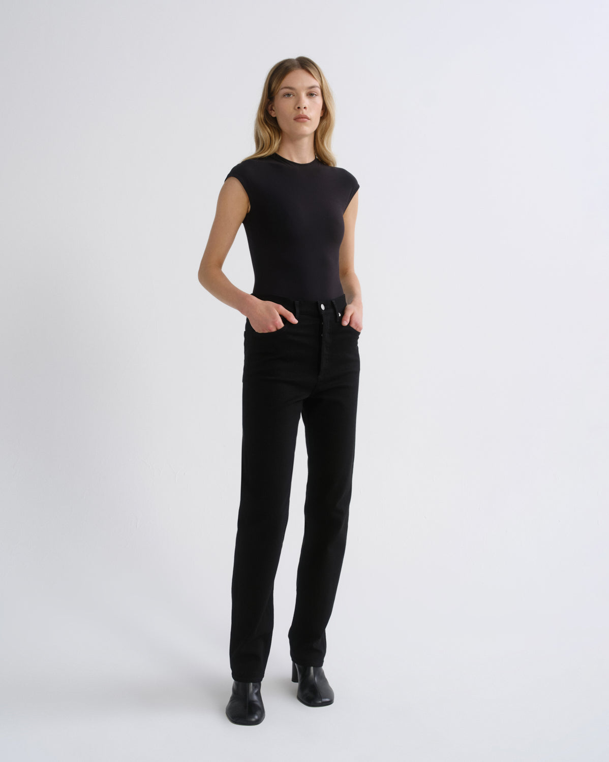 A woman modeling a black short-sleeve crewneck top and black high-waisted straight-leg jeans with black leather ankle boots, standing against a light gray studio background.