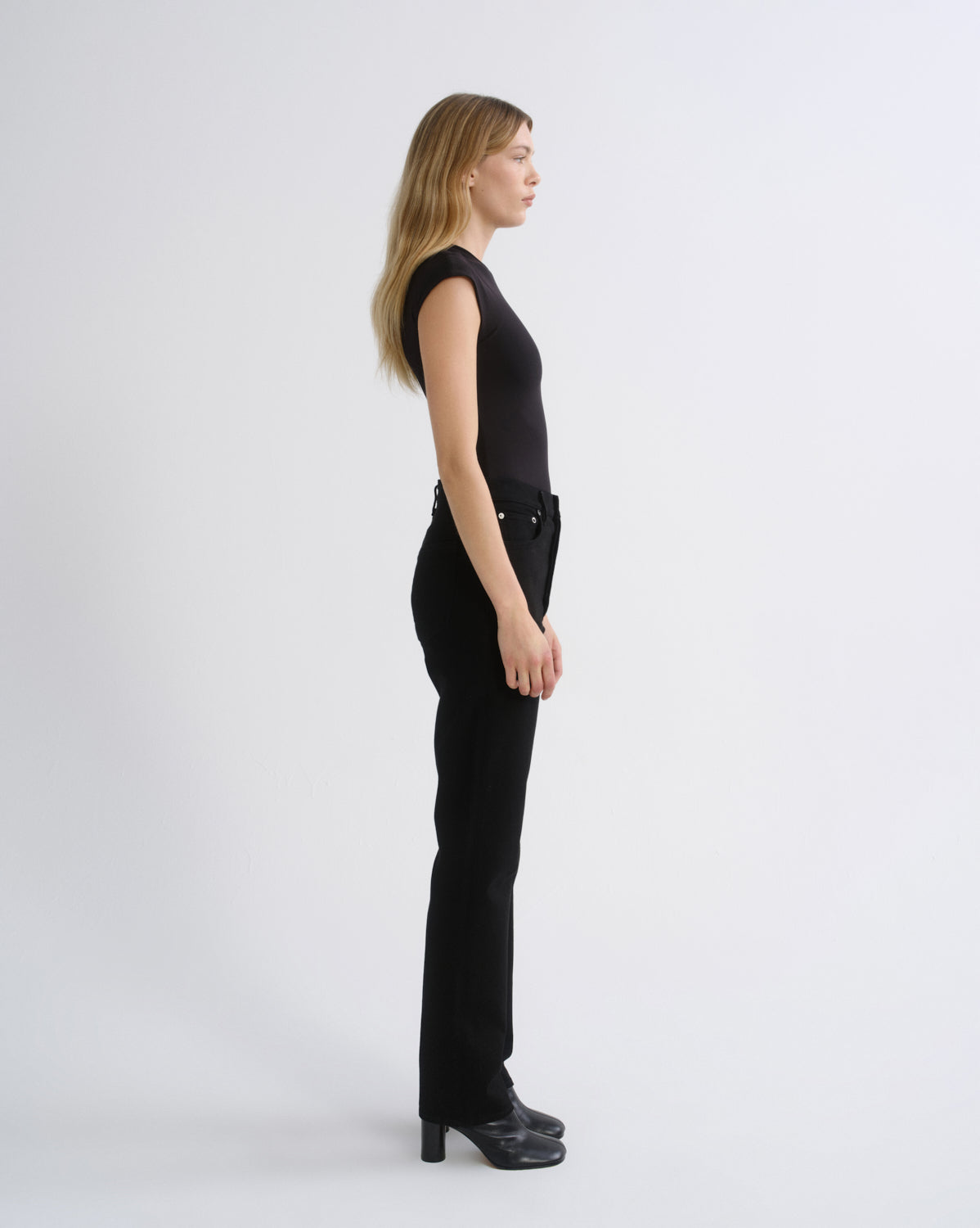 A woman in a black sleeveless top and black high-waisted flared trousers with black leather boots, shown in profile against a light gray studio background.