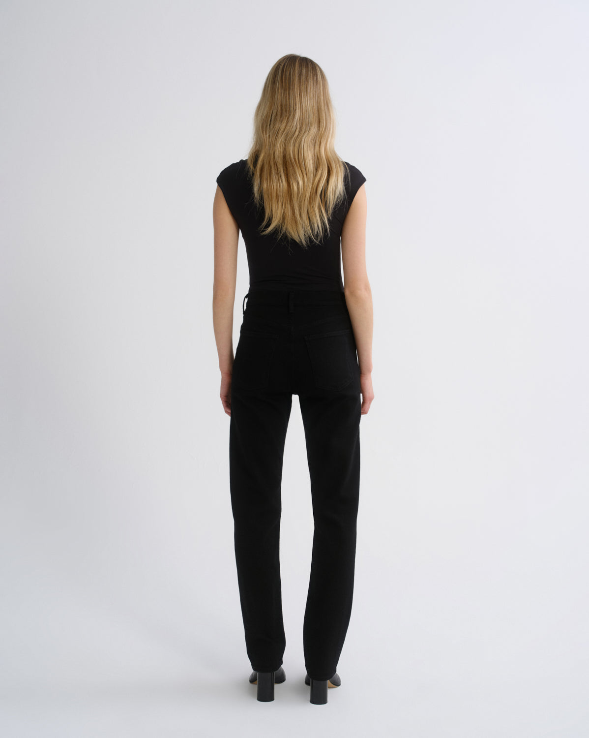 A woman with long blonde hair is shown from the back, wearing a black sleeveless top and black pants against a plain light gray studio background.