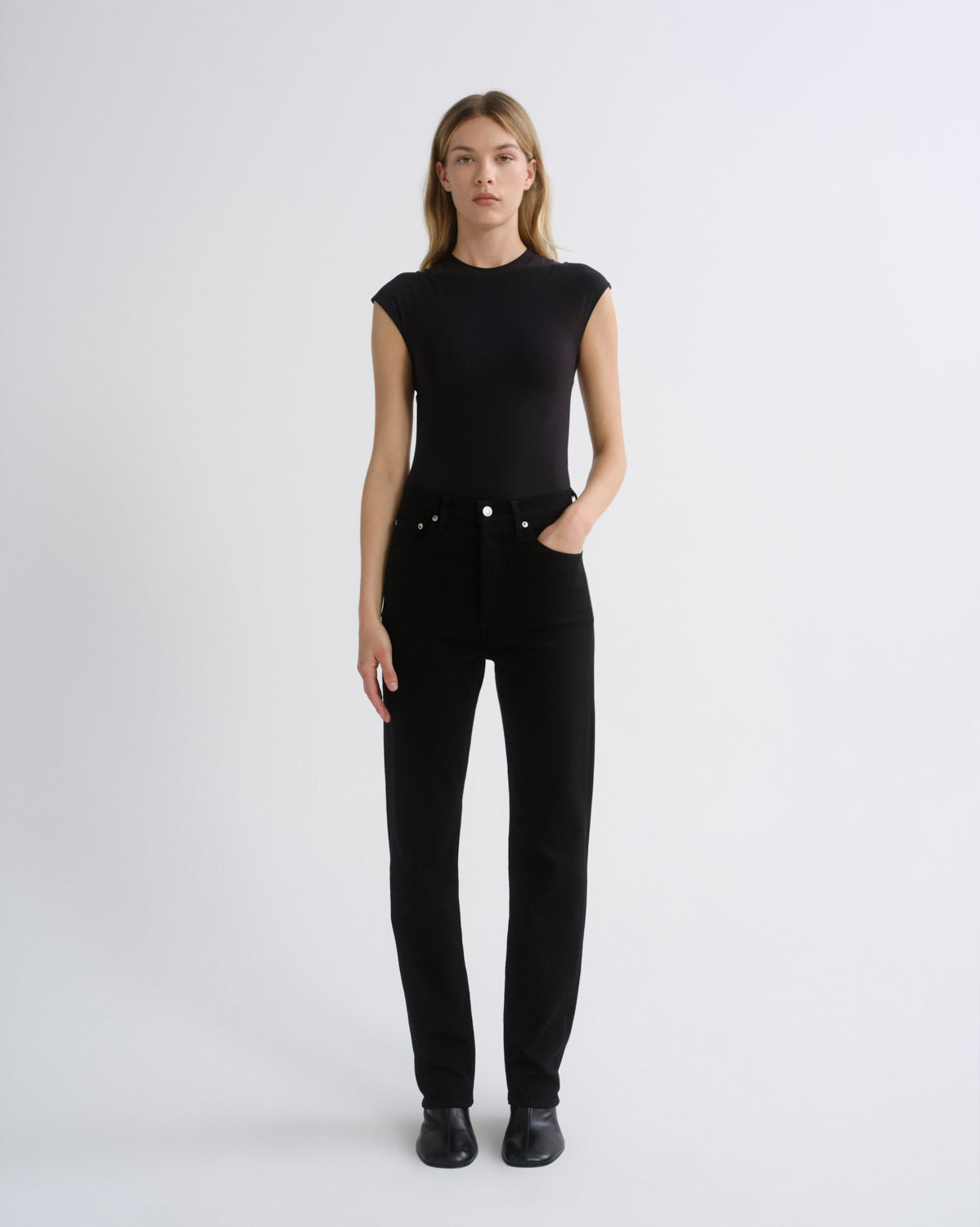 A woman modeling a black sleeveless mock-neck top and black straight-leg jeans against a plain light gray studio background.