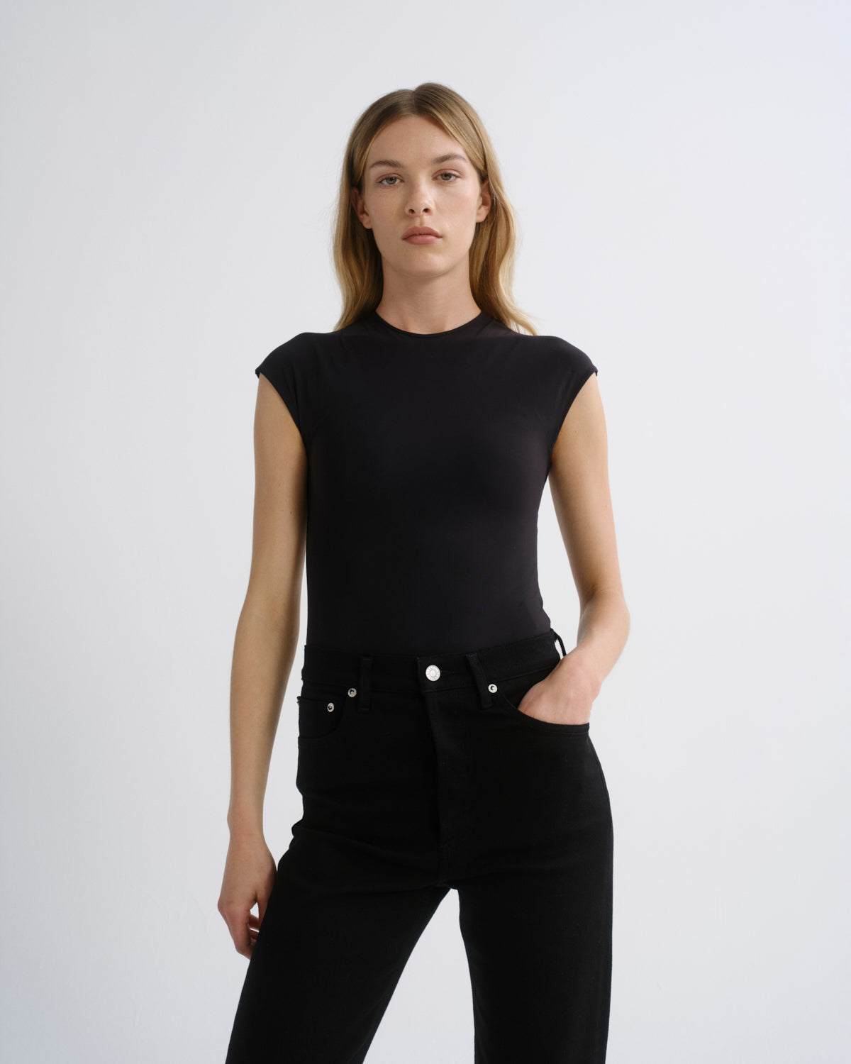 A woman modeling a black short-sleeve crewneck top paired with black jeans against a light gray studio background.