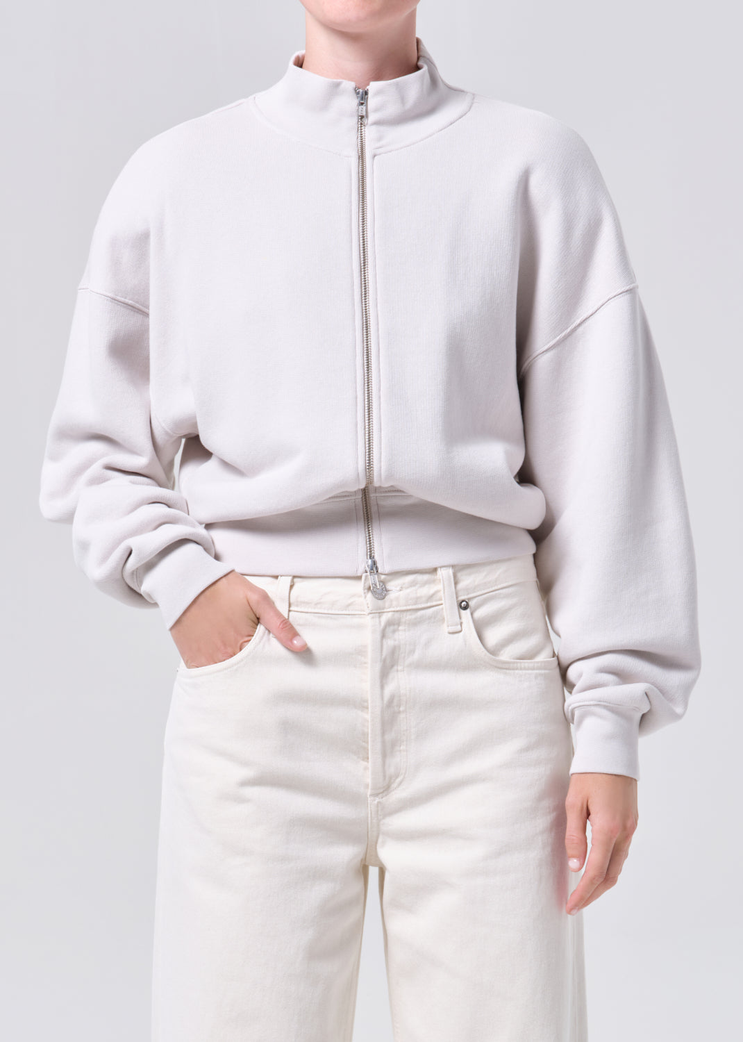Person wearing a light gray zip-up sweatshirt and white pants on a plain background