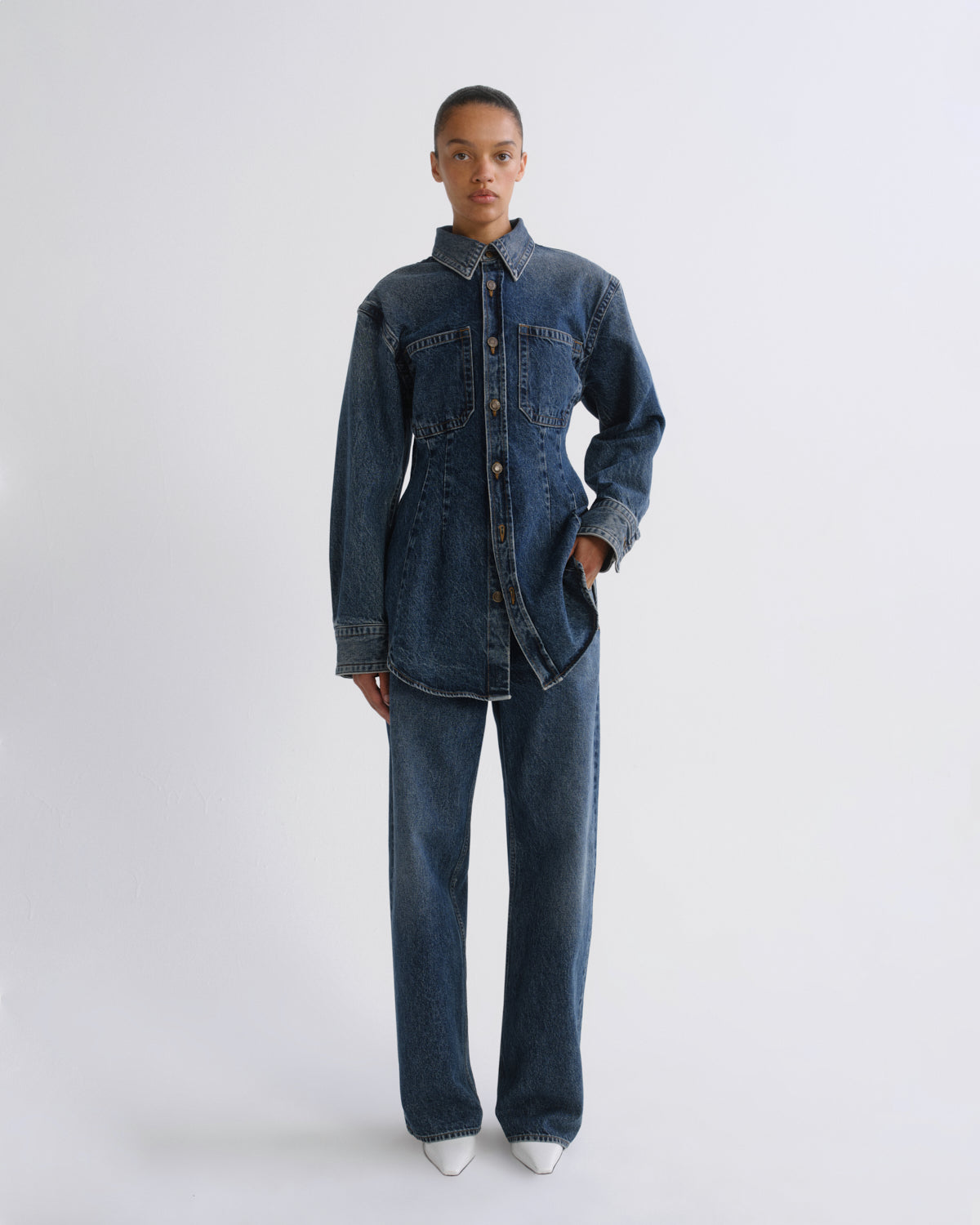 A model stands against a light gray studio background wearing a dark blue denim shirt-jacket and matching wide-leg jeans, paired with white pointed shoes.
