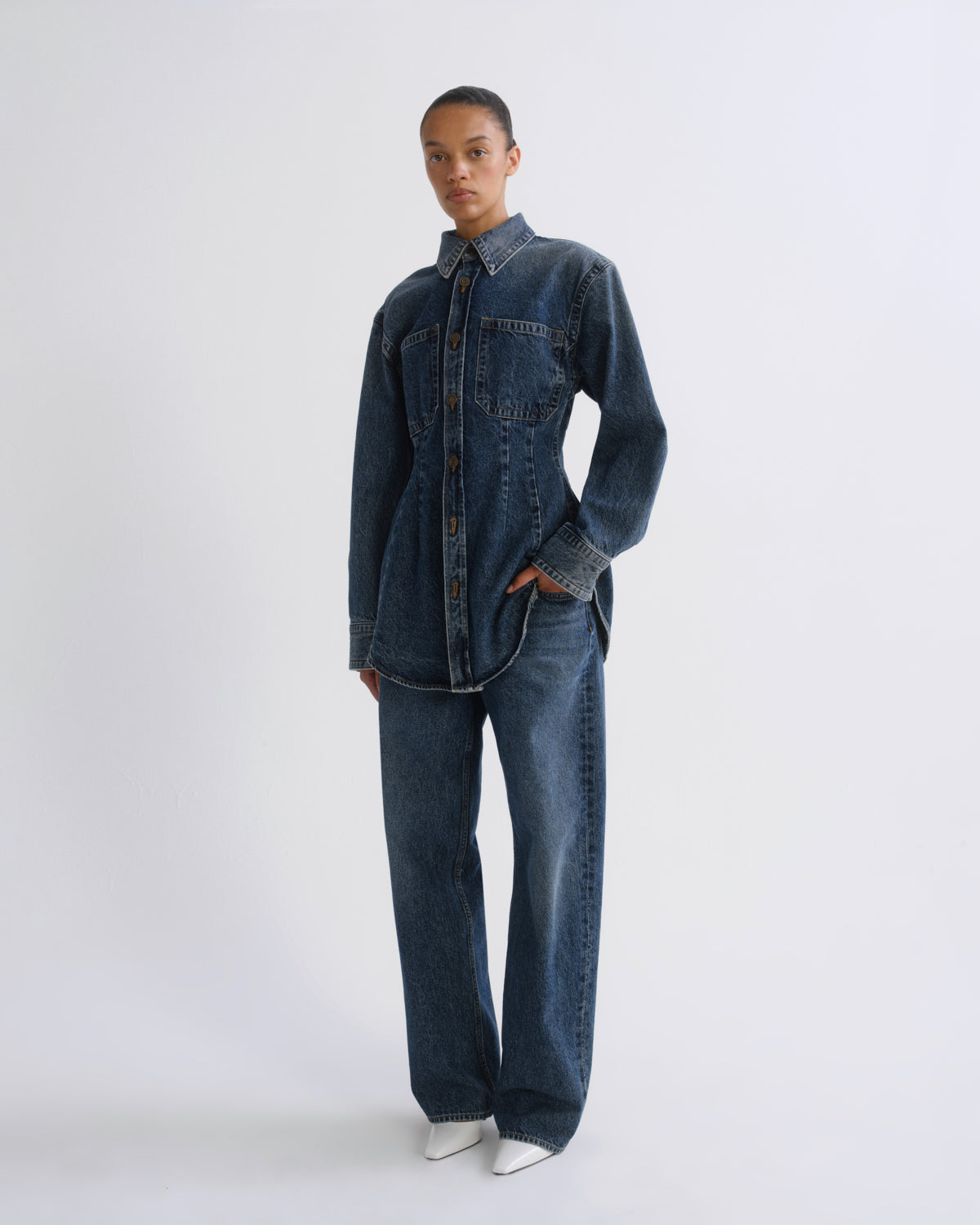 A model wearing a dark blue denim shirt and matching wide-leg jeans, standing against a plain light gray studio background.