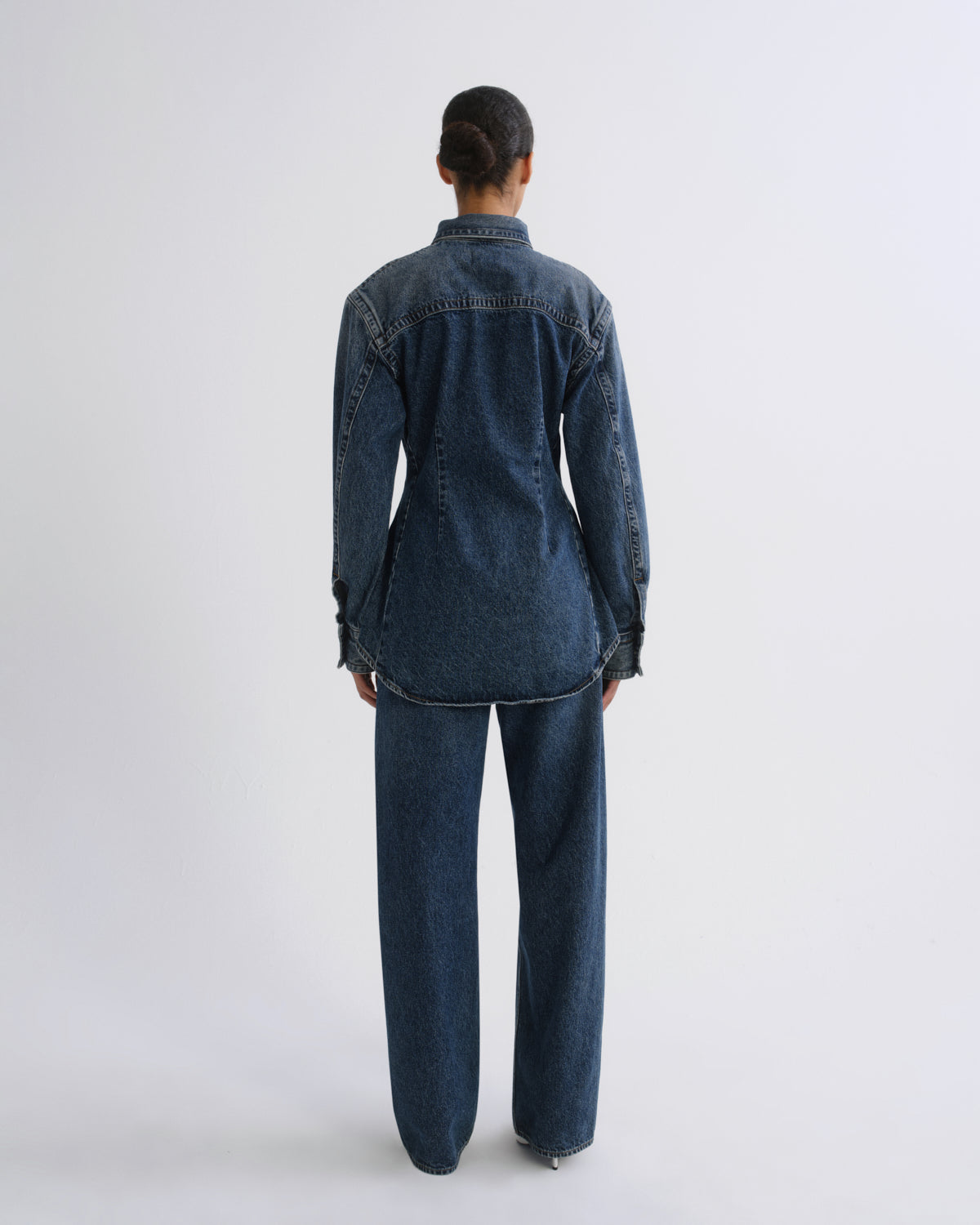A model viewed from the back wearing a dark blue denim shirt-jacket and matching wide-leg jeans against a light gray studio background.