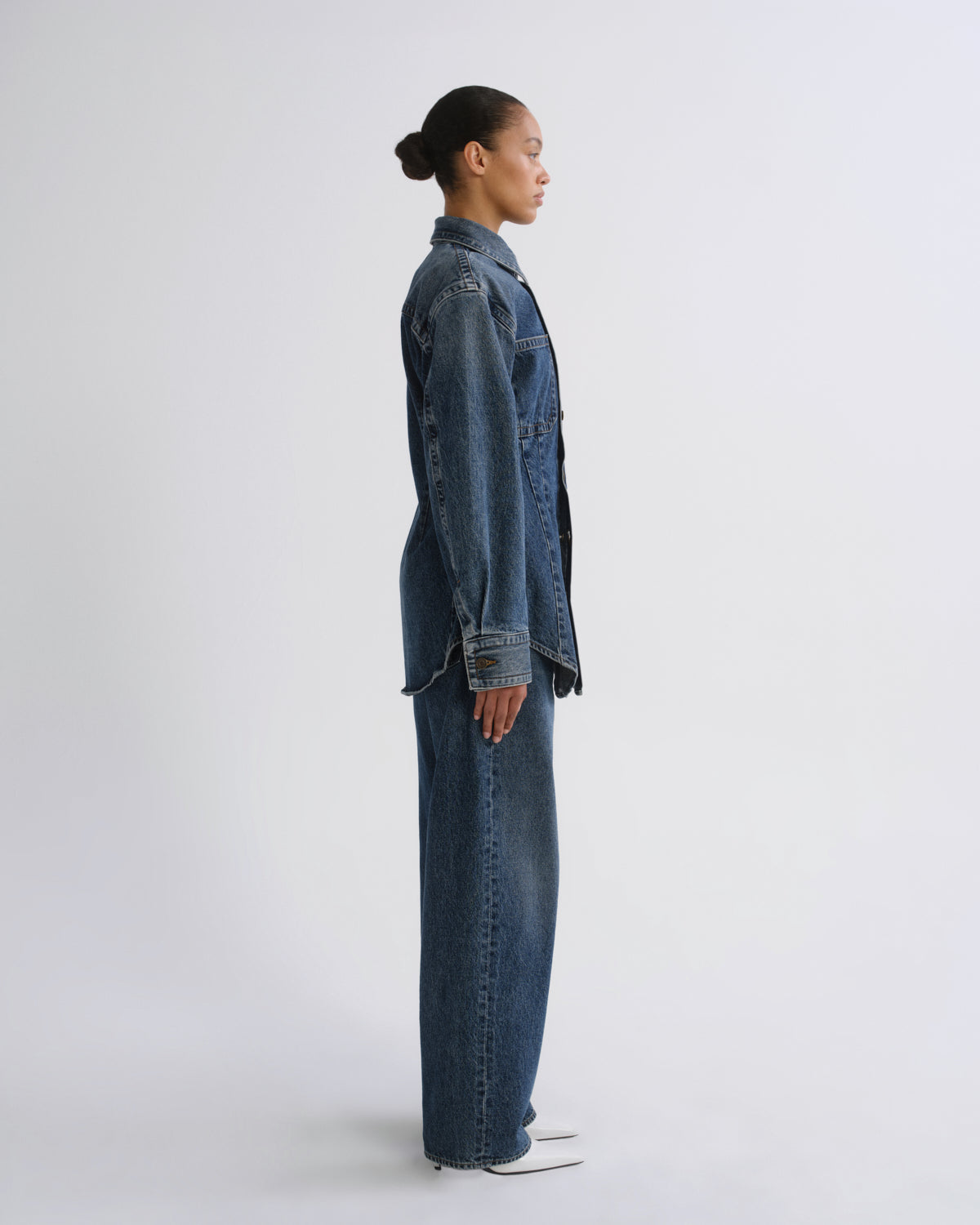 A model in profile wearing a matching blue denim jacket and wide-leg denim pants, standing against a plain light gray studio background.
