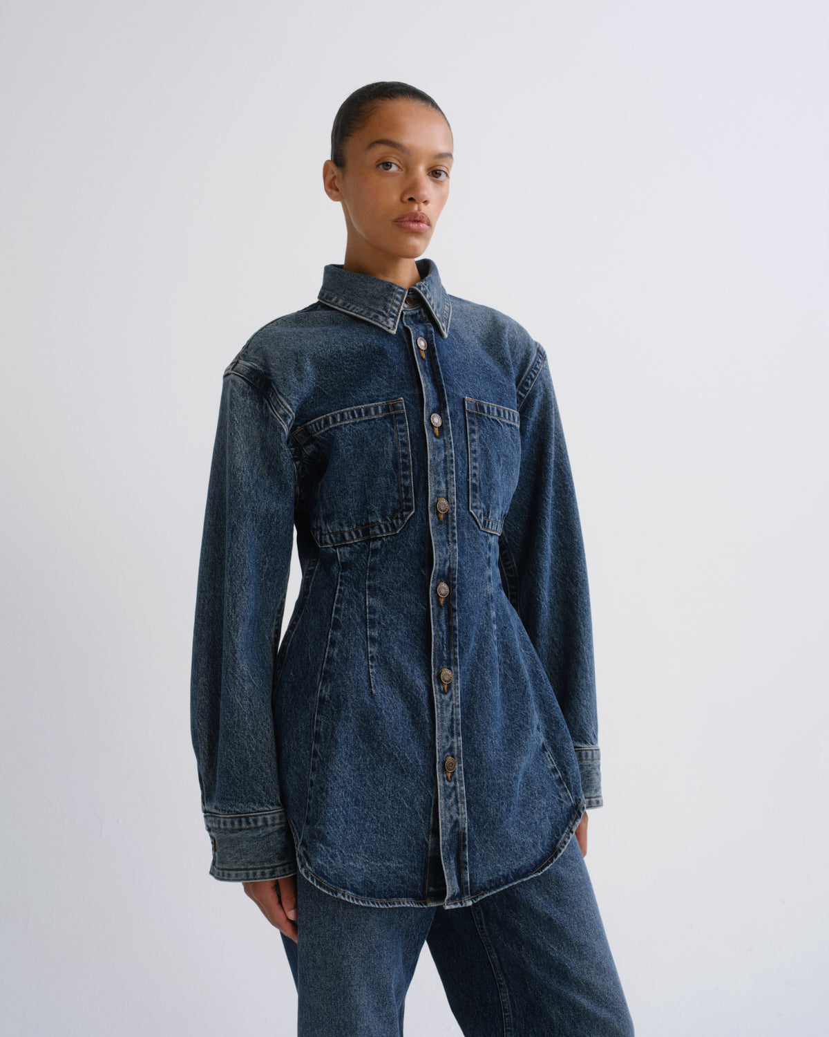 A model wearing a dark blue denim button-up shirt jacket with front chest pockets and matching dark blue denim trousers, photographed against a plain light gray studio background.