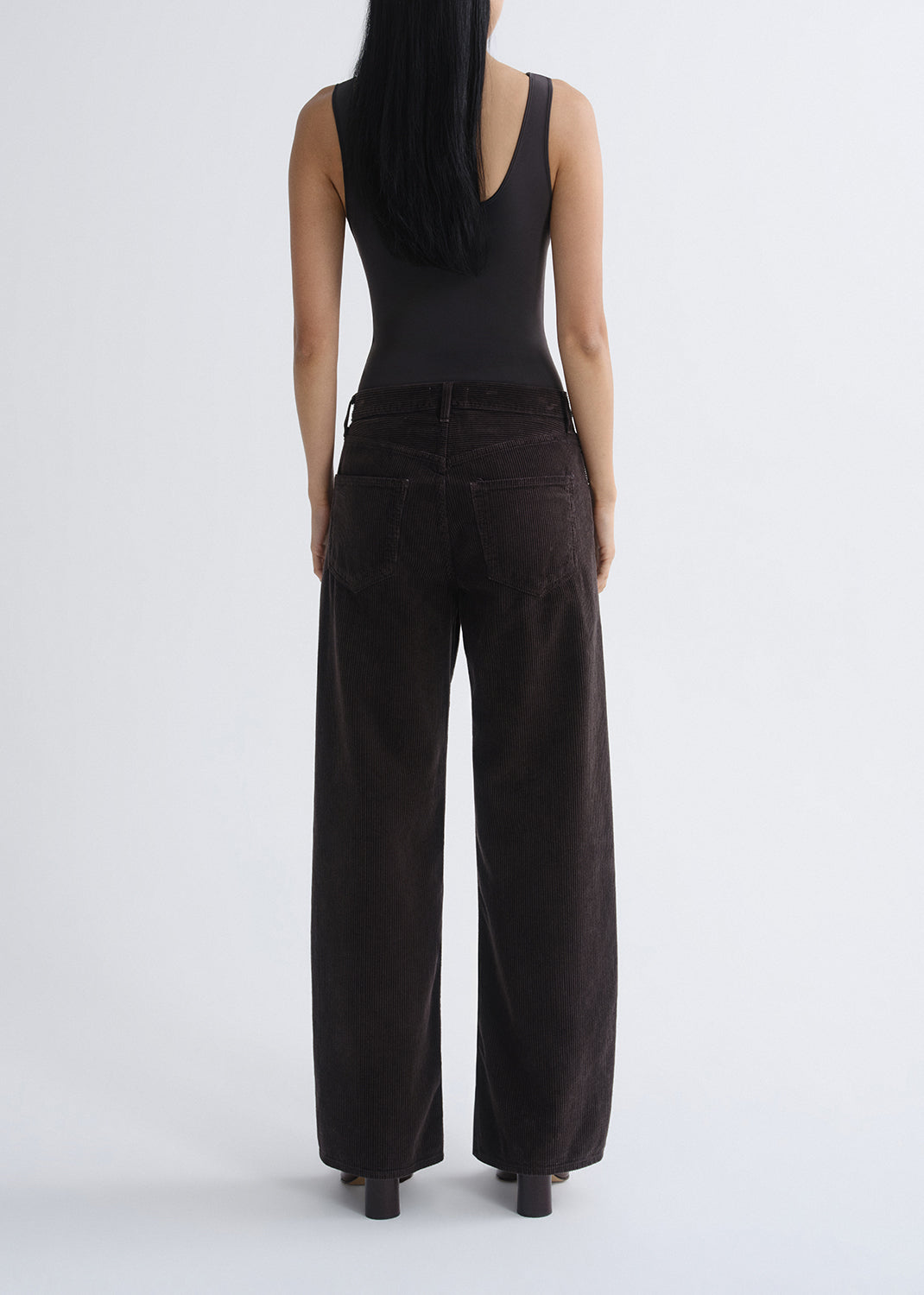 Person wearing a black tank top with black wide leg jeans viewed from the back on a grey background