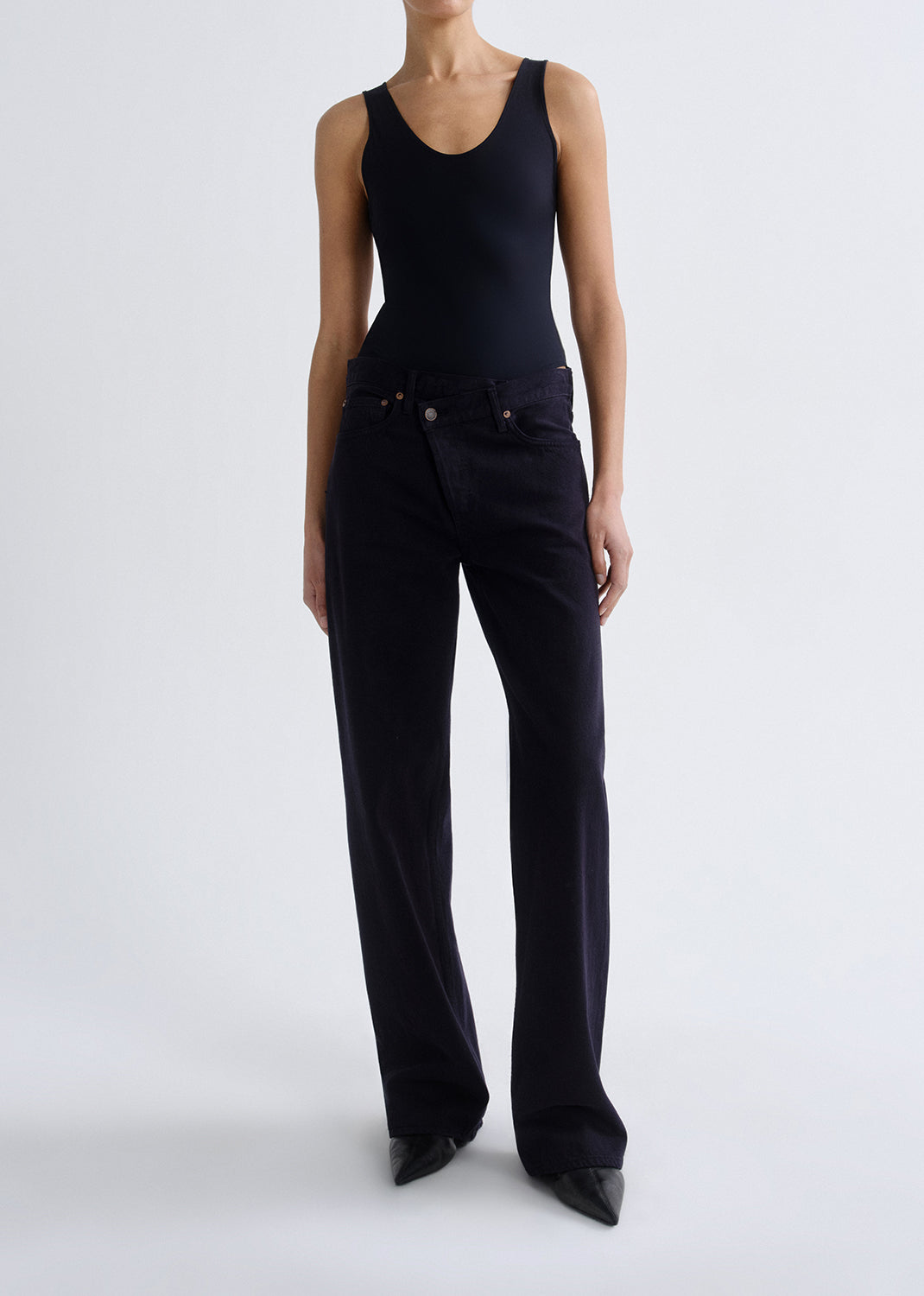 Person wearing a black tank top with black straight leg jeans on a grey background.