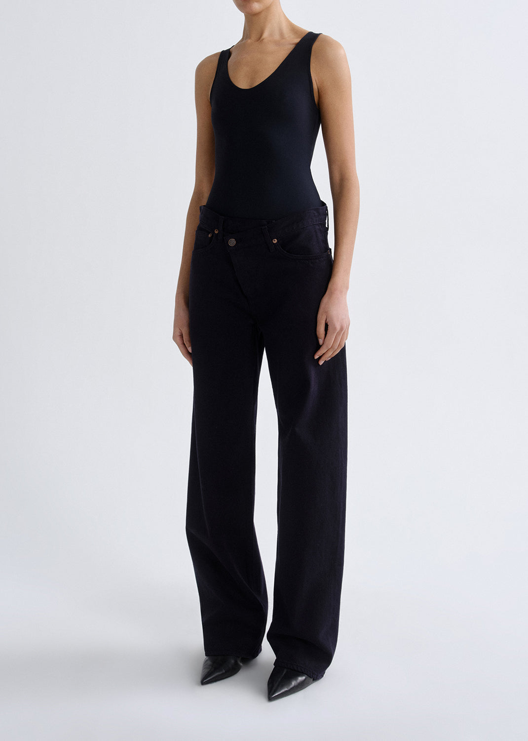 Person wearing a black tank top with black straight leg jeans on a grey background.