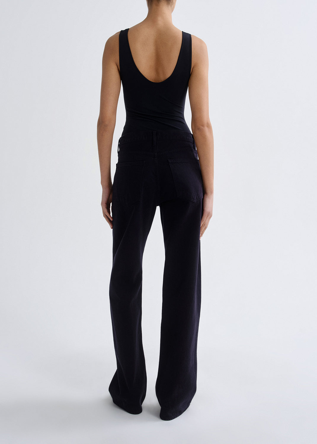 Person wearing a black tank top with black straight leg jeans viewed from the back on a grey background.