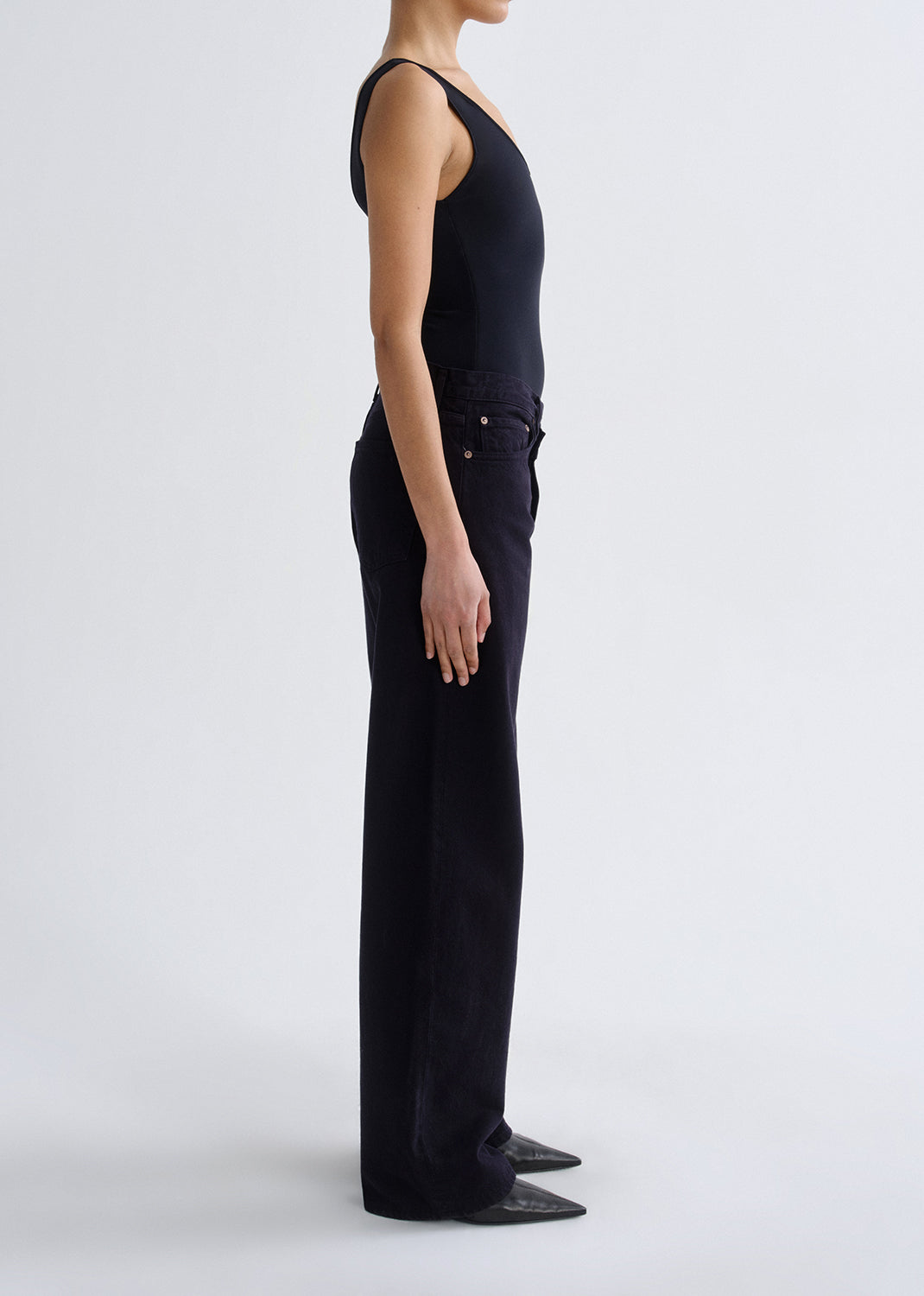 Person wearing a black tank top with black straight leg jeans viewed from the side on a grey background.