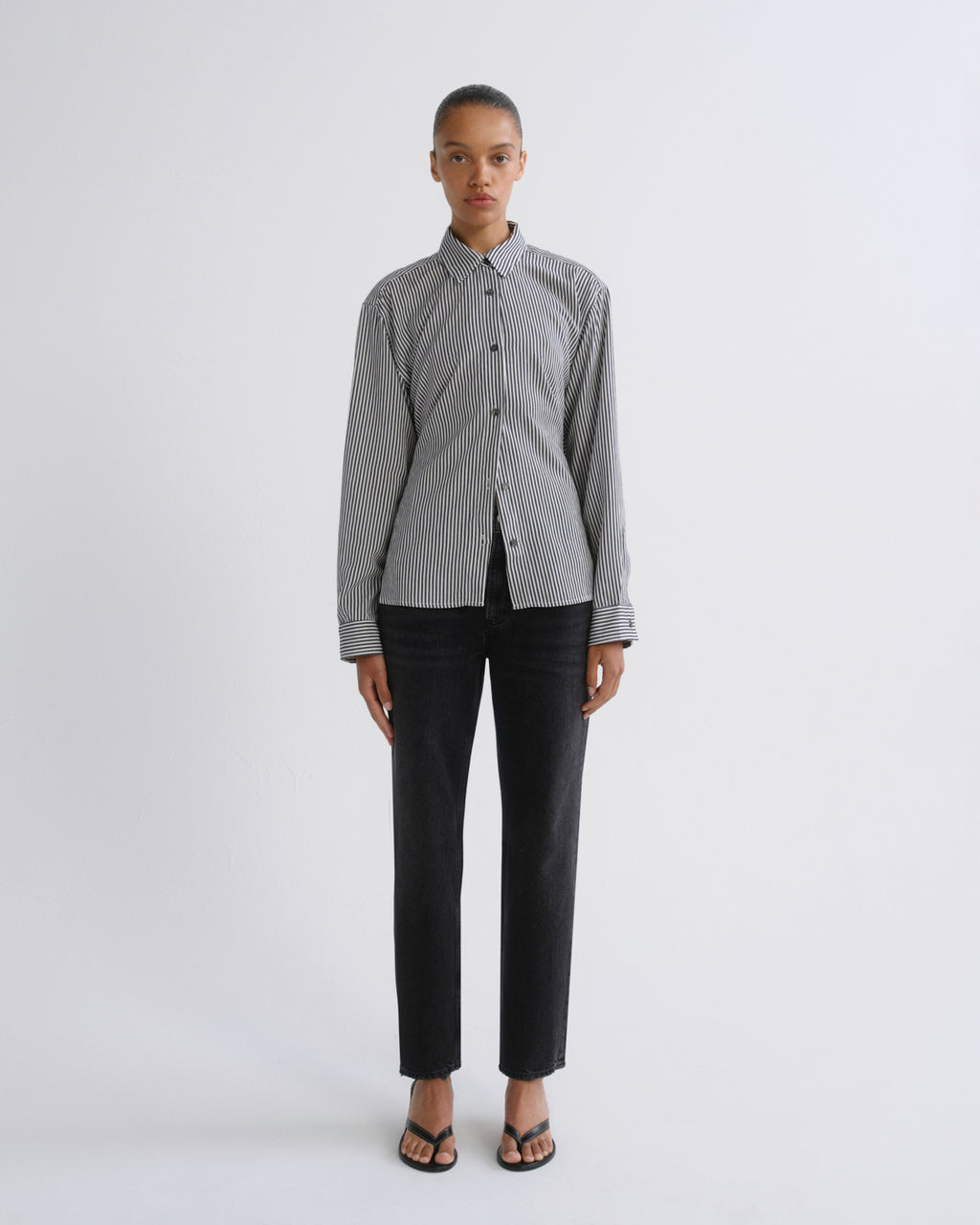 A model stands against a light gray studio background wearing a gray and white vertically striped button-down shirt, black trousers, and black sandals.