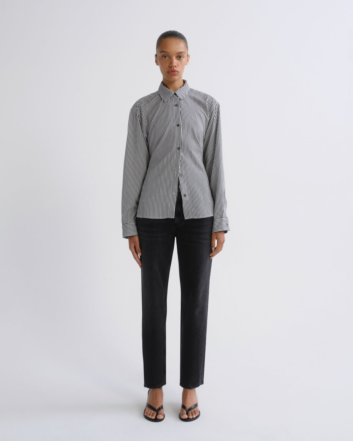 A model stands against a light gray studio background wearing a gray and white vertically striped button-down shirt, black trousers, and black sandals.
