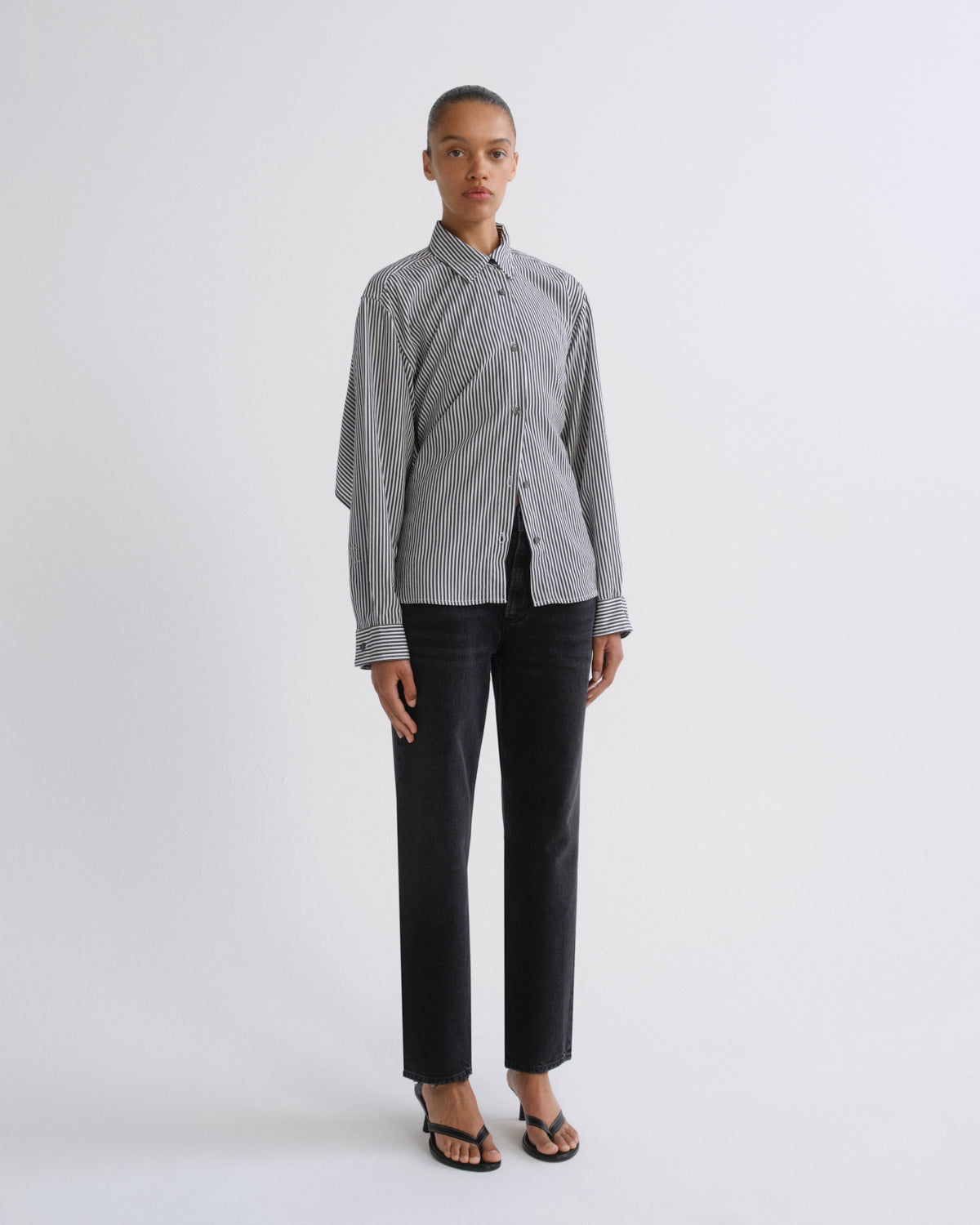 A model stands against a light gray studio background wearing a gray and white vertically striped button-up shirt with textured sleeve detailing, paired with black trousers and black sandals.