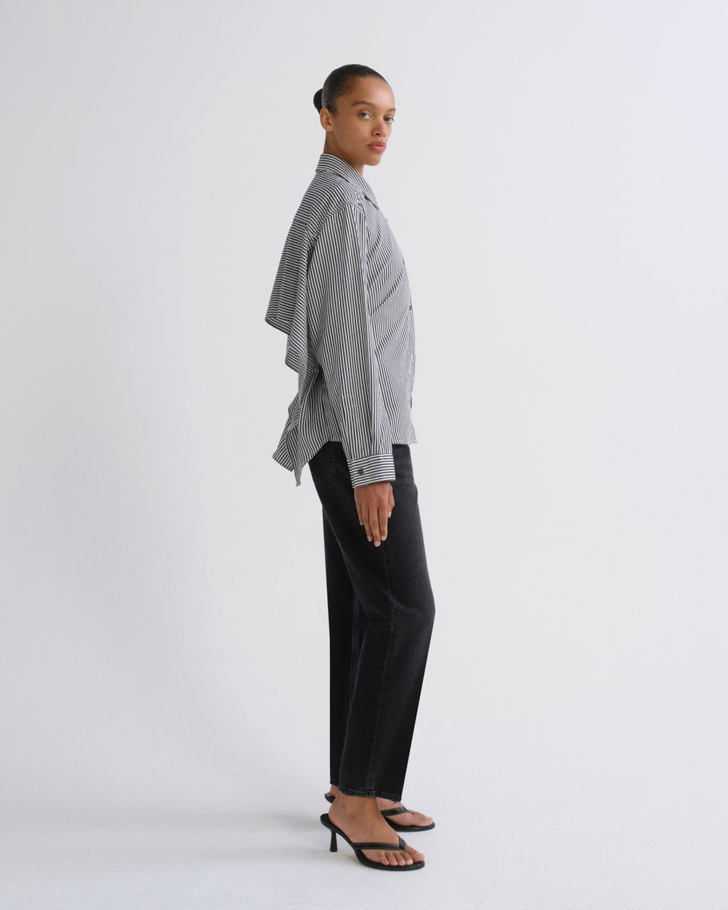 A model is shown in profile wearing a light gray and white vertically striped button-down shirt with draped sleeves, paired with black wide-leg trousers and black heeled sandals, against a plain light gray studio background.