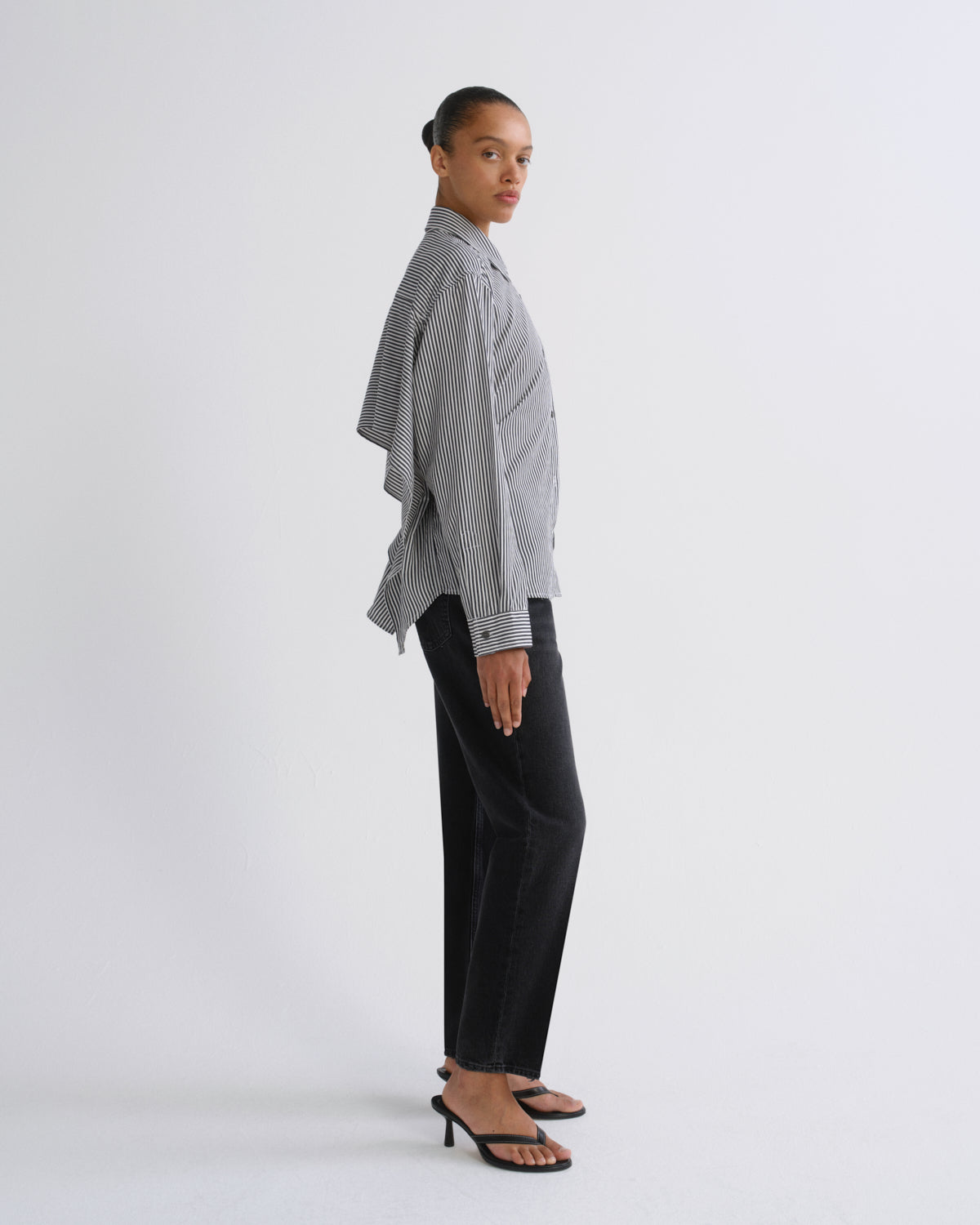 A model is shown in profile wearing a light gray and white vertically striped button-down shirt with draped sleeves, paired with black wide-leg trousers and black heeled sandals, against a plain light gray studio background.