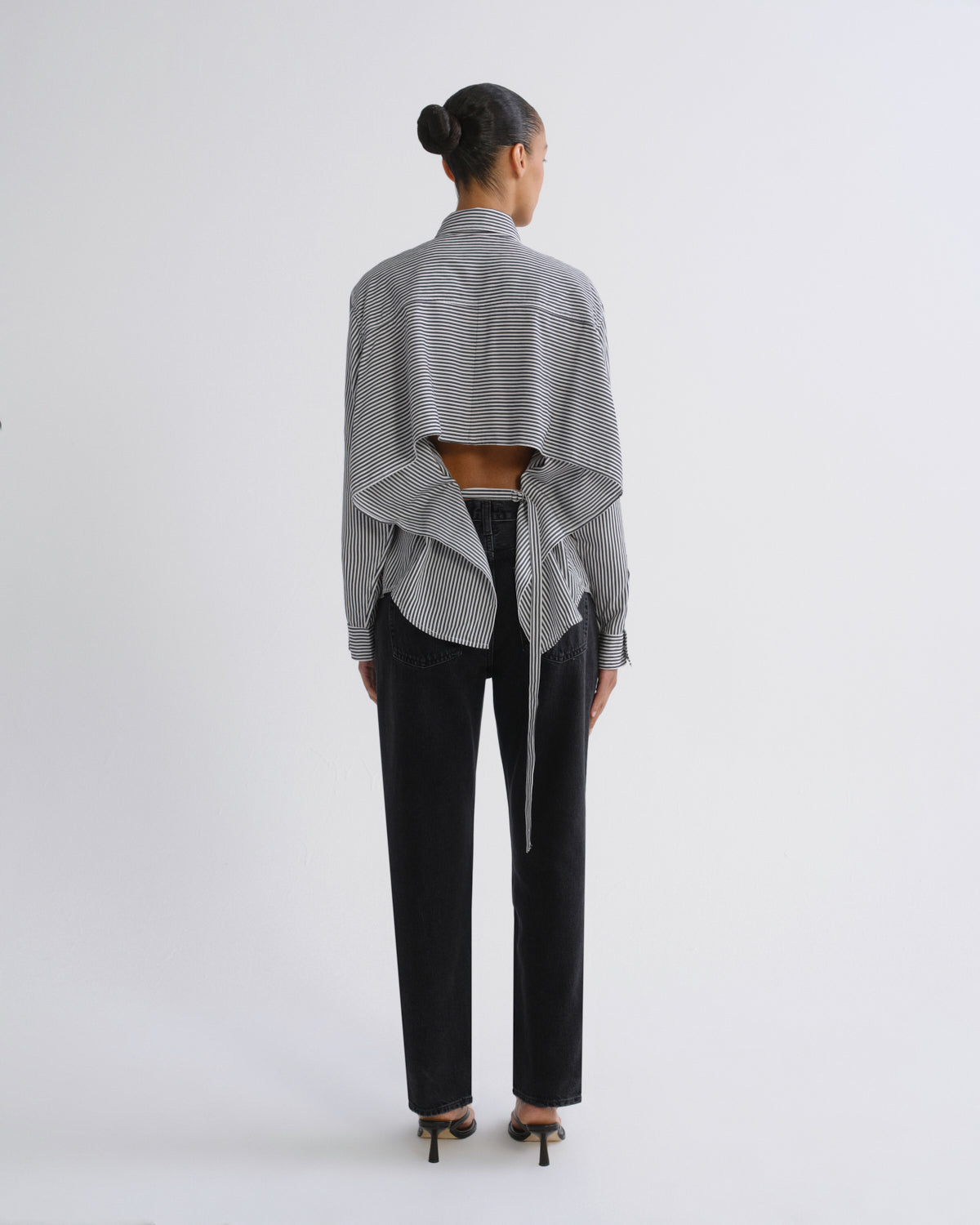 A model viewed from the back wearing a gray and white striped cropped shirt with a dramatic draped ruffle design, paired with black pants and black heels, against a plain white studio background.