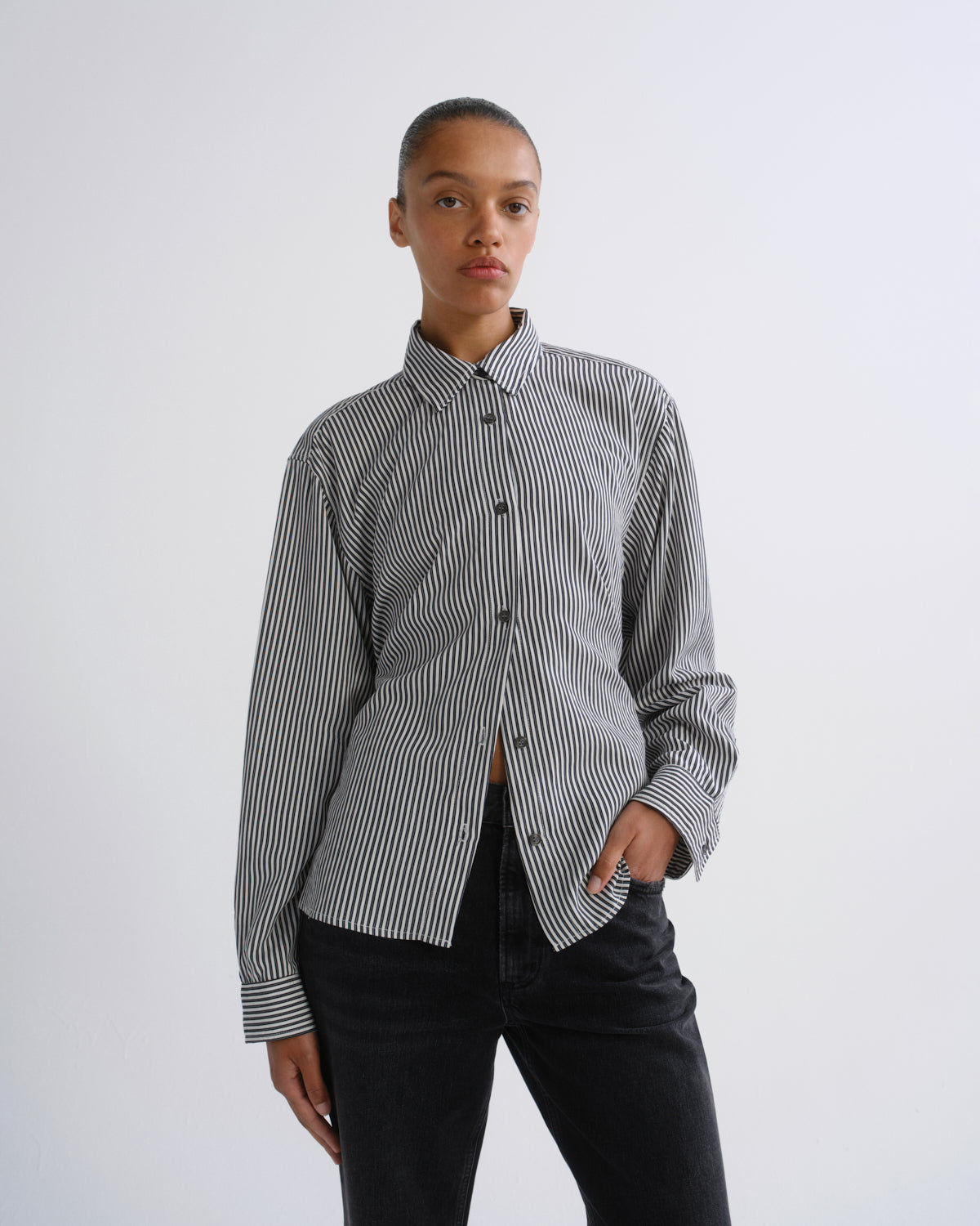 A model wearing a black and white vertically striped button-down shirt with black jeans, standing against a plain light gray studio background.