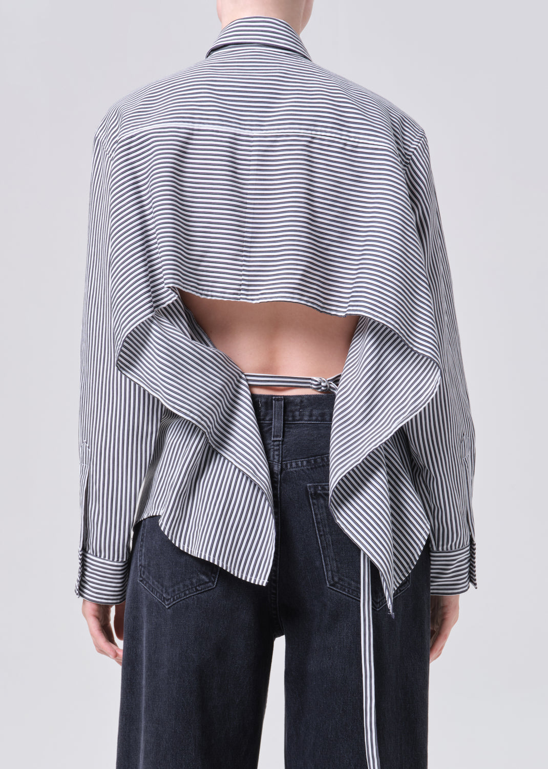 Person wearing a black and white striped shirt with a draped design over dark pants on a light gray background