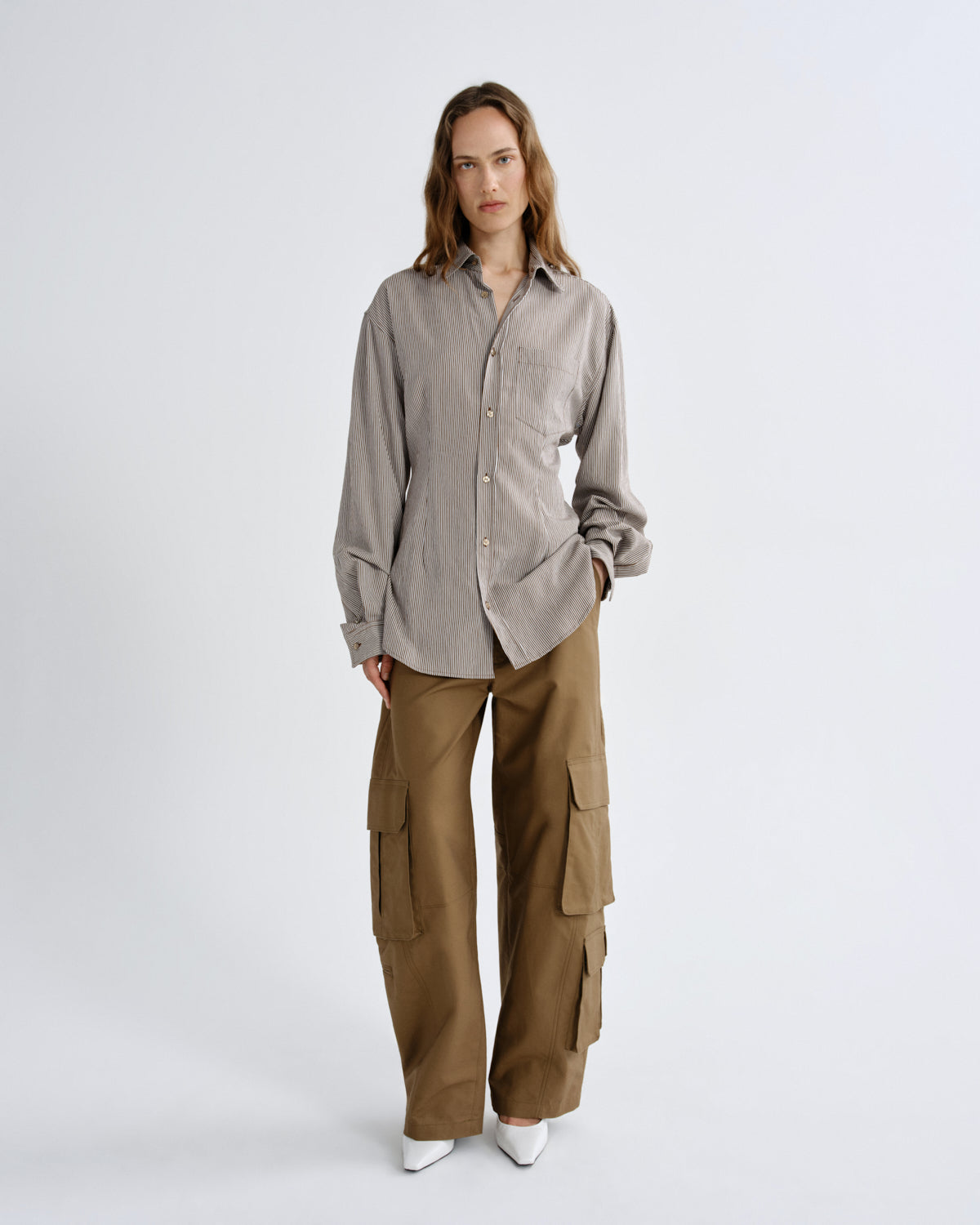 A model stands against a light gray studio background wearing a light taupe-gray long-sleeve button-up shirt and wide-leg brown cargo pants.