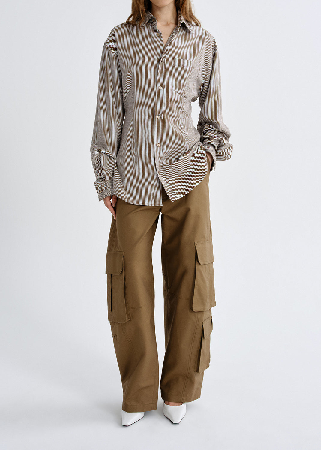 A model wearing a light taupe long-sleeve button-up shirt and wide-leg brown cargo pants, paired with white pointed-toe heels, against a grey background.