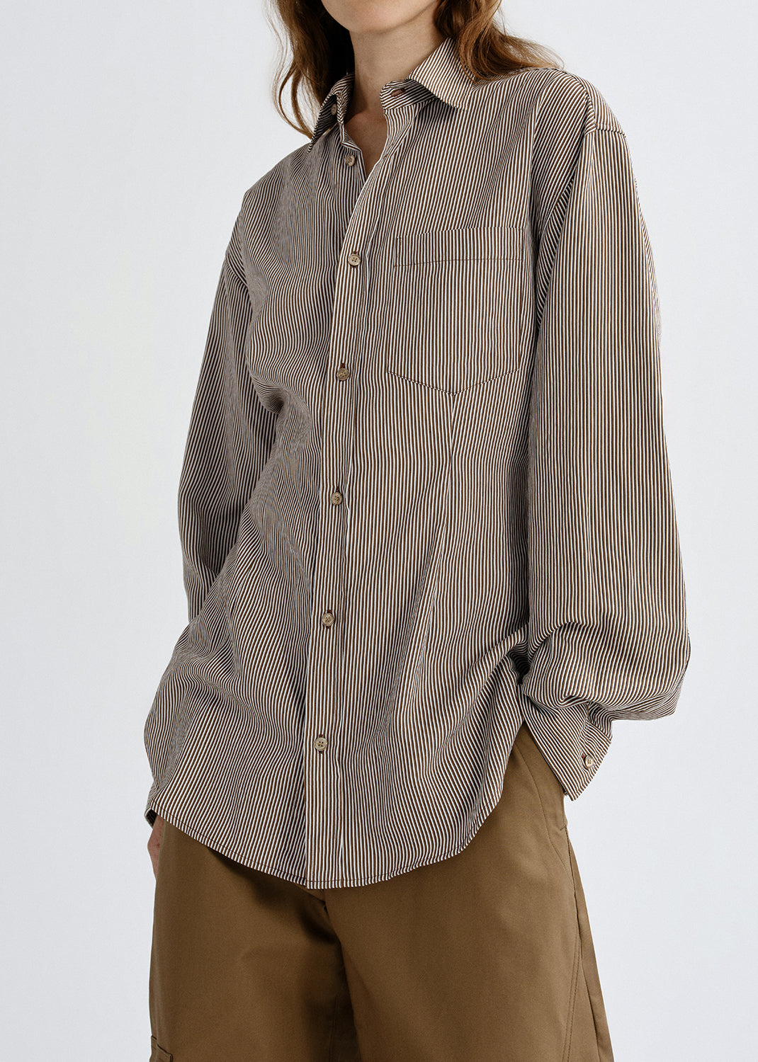 A woman wearing an oversized long-sleeve button-up shirt with fine vertical stripes in taupe and brown, paired with brown pants, photographed against a grey background.