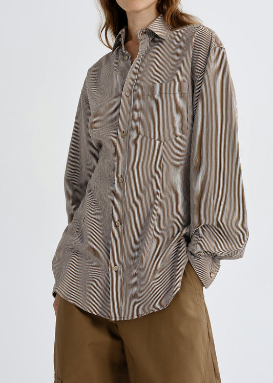 A woman wearing an oversized long-sleeve button-up shirt with fine vertical stripes in taupe and brown, paired with brown pants, photographed against a grey background.