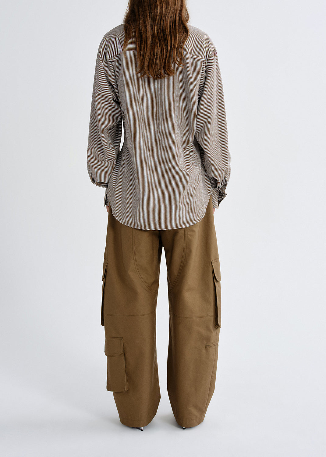 A model shown from the back wearing a light taupe-gray long-sleeve linen shirt and wide-leg brown cargo pants, set against a grey studio background.
