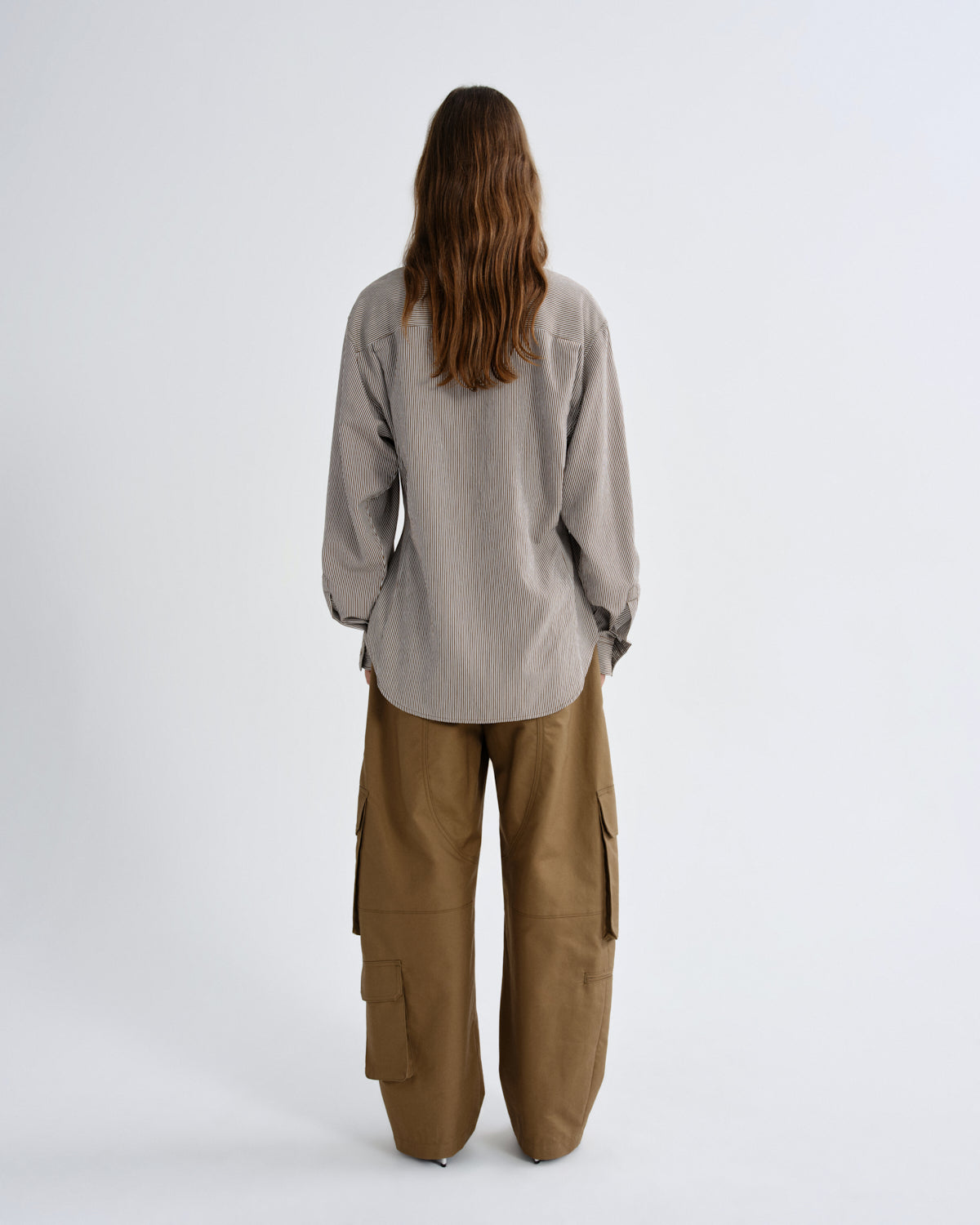 A model viewed from the back wearing a light gray oversized long-sleeve shirt and wide-leg brown cargo pants, standing against a plain light gray studio background.