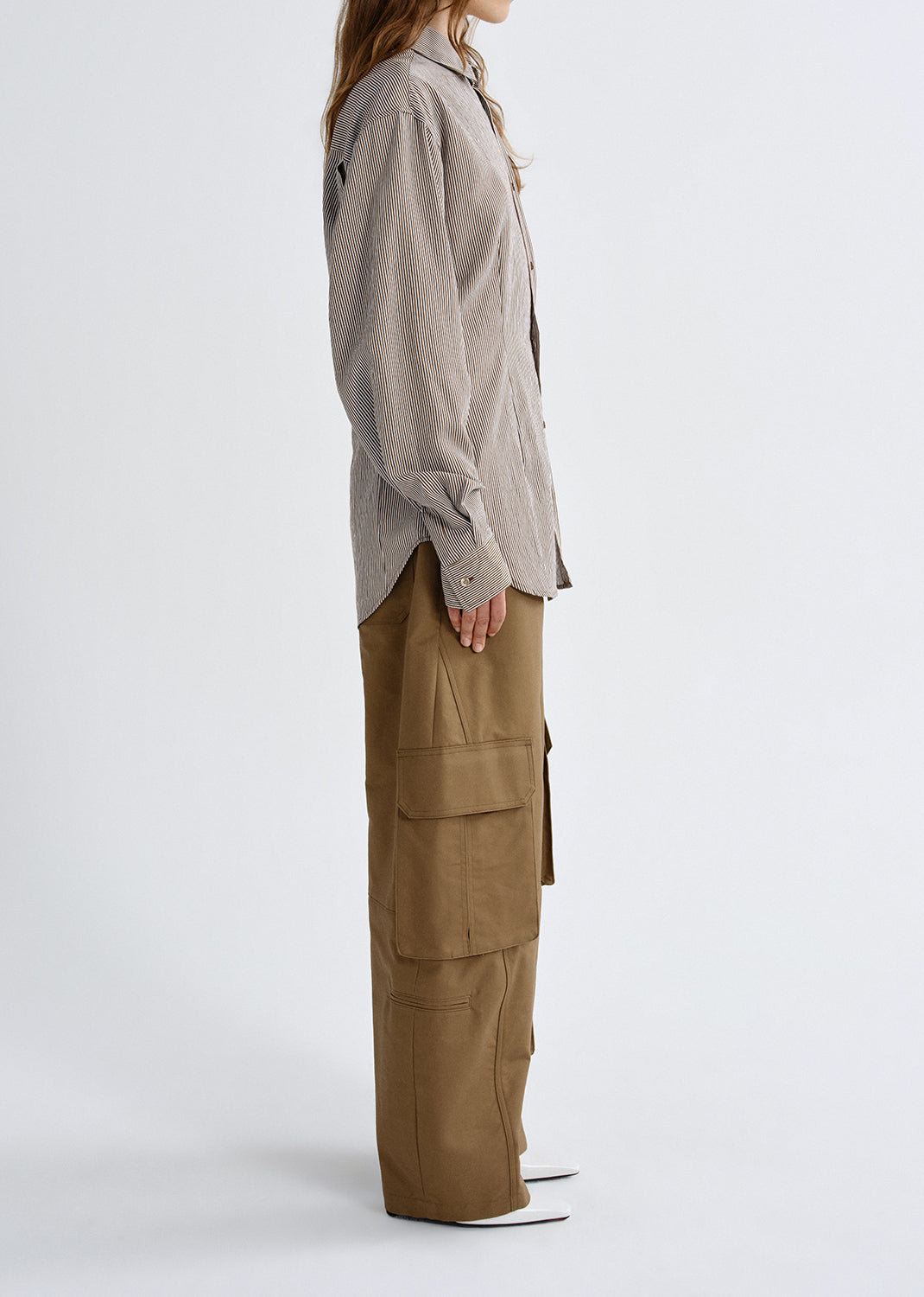 A side-profile studio shot of a model wearing a light taupe linen button-up shirt and wide-leg brown cargo pants against a grey background.