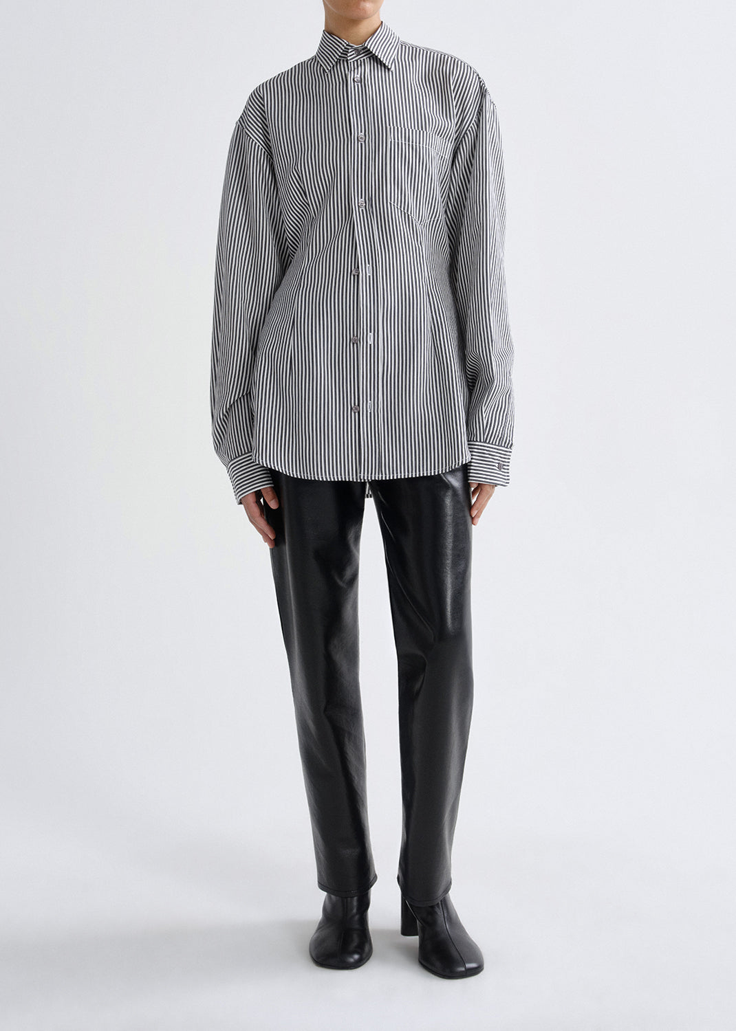 A model wearing a black and white vertically striped long-sleeve button-up shirt, paired with black leather trousers and black boots, standing against a grey background.